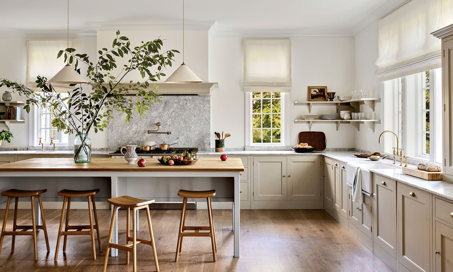 A bright, airy kitchen with white walls features a large island with wooden stools, pendant lights, and a vase with green branches. Sunlight streams in through the windows.