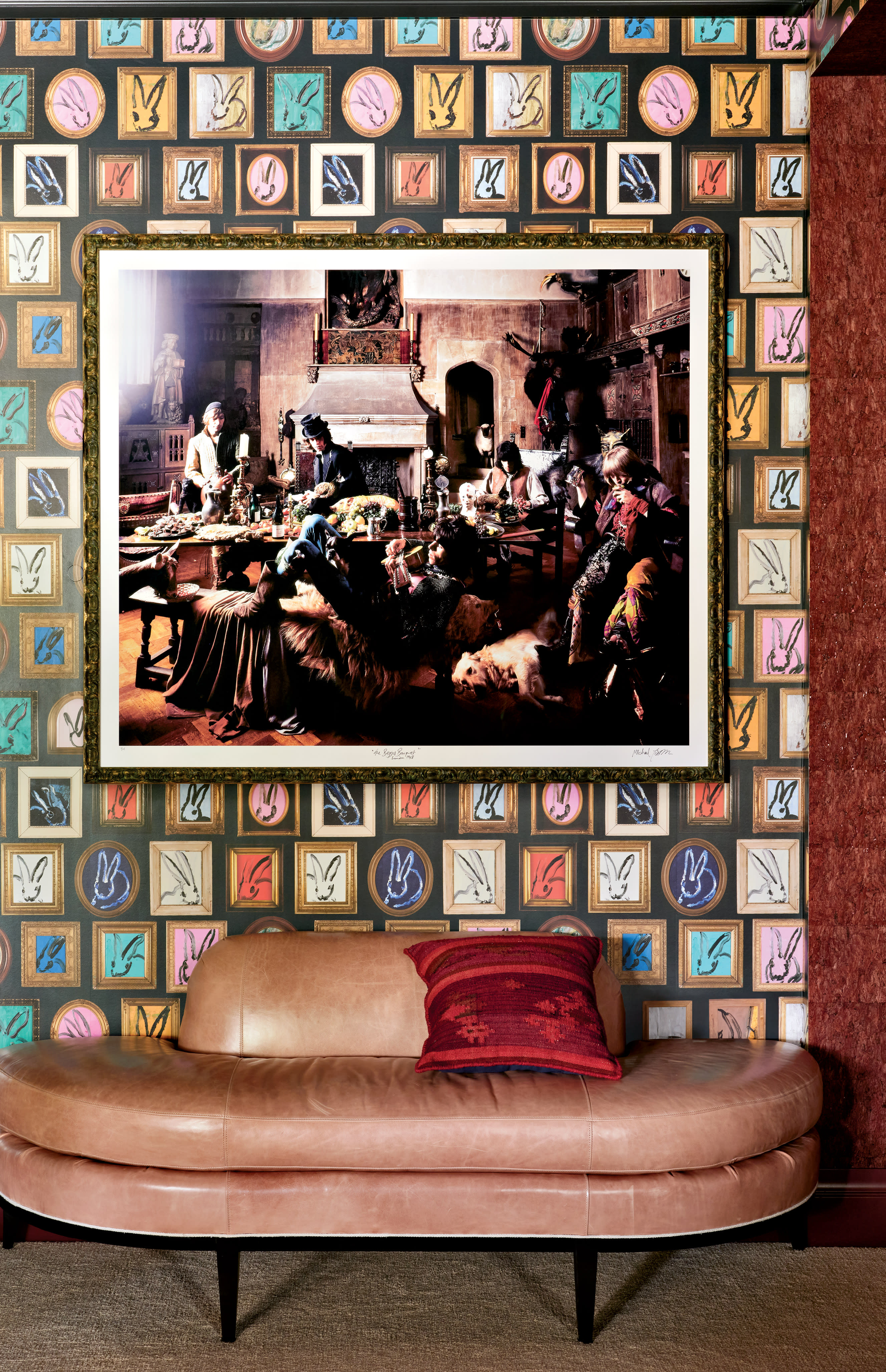 A stylish living room features a painting centered on a vibrant wall with colorful, patterned wallpaper. Below, a curved beige sofa with a red pillow adds to the modern decor.