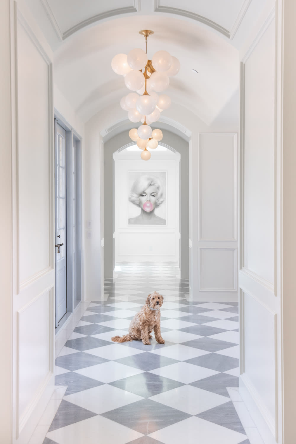 A dog sitting on a checkered floor in an elegant hallway with white walls and gold lighting.