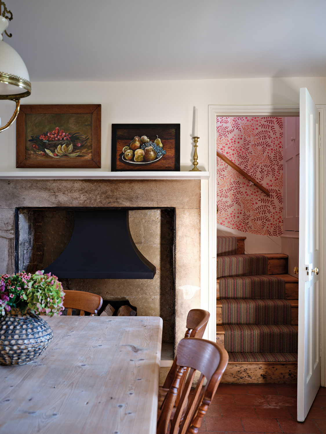 A dining room with a wooden table and chair facing a large stone-surround fireplace, with an open door revealing a staircase with patterned wallpaper.