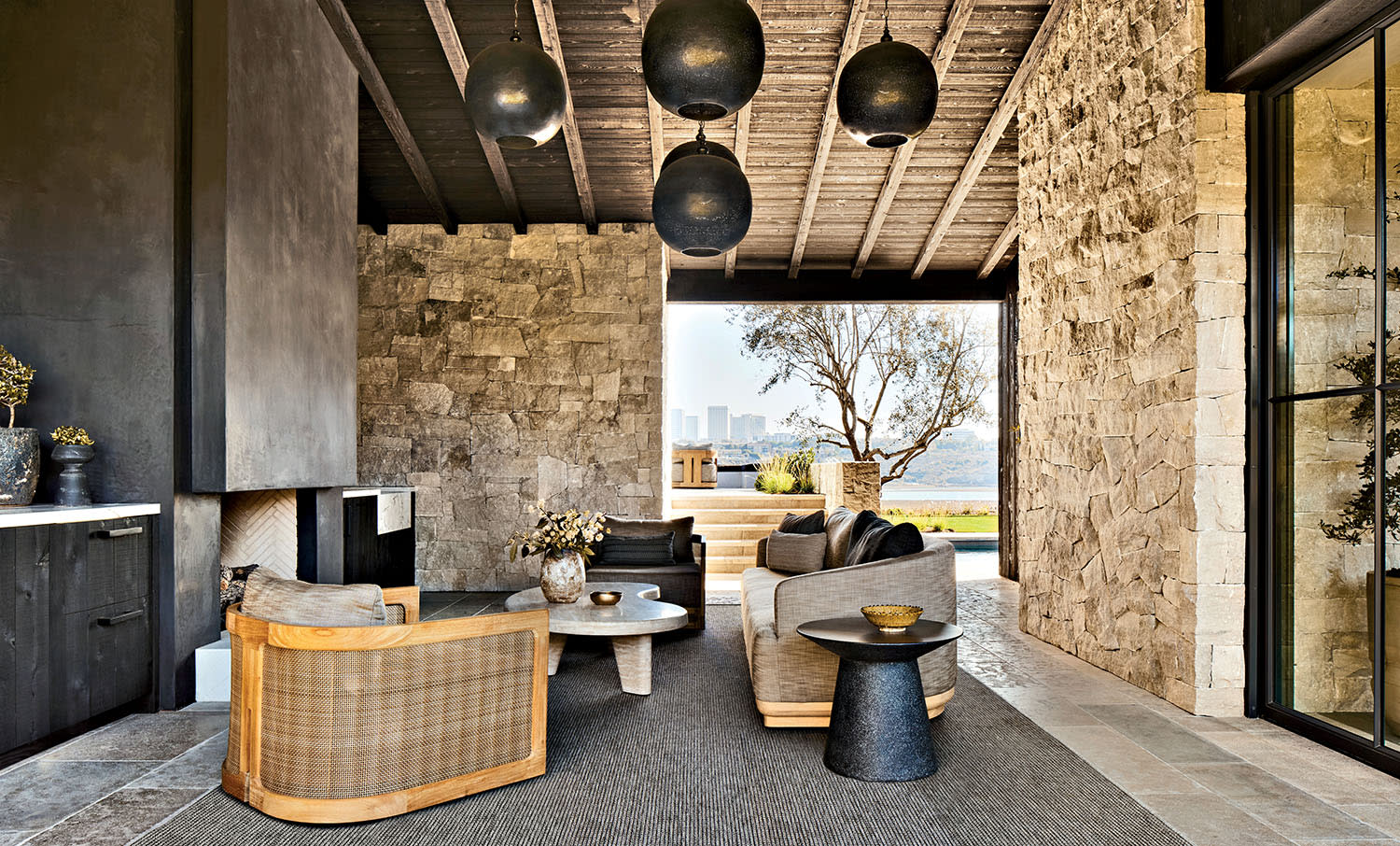 Spacious modern patio with stone walls, dark ceiling, and black pendant lights. Features light wooden furniture, a gray rug, and a scenic outdoor view.