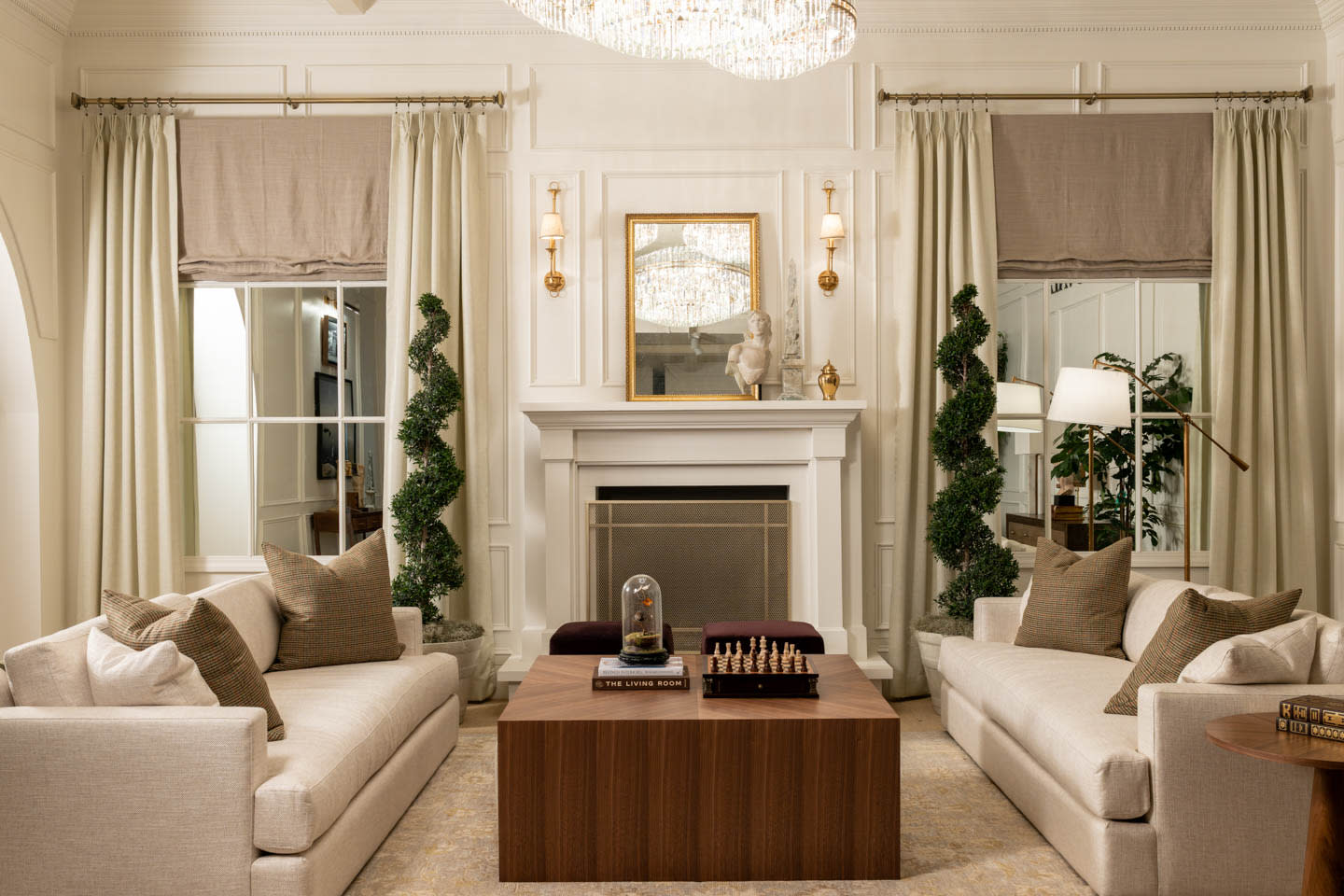 Elegant living room with a symmetrical design features beige sofas, a wooden coffee table with a chessboard, and a fireplace with a statue. 