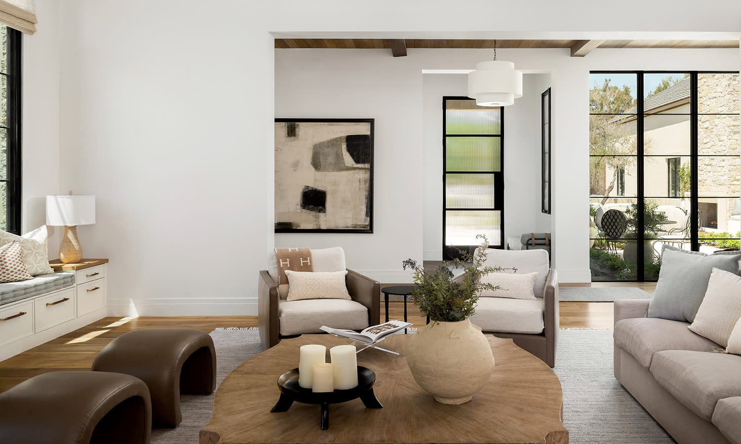 Modern living room with neutral tones. A beige sofa, abstract art, vase on wooden table, and large windows create an open, inviting atmosphere.