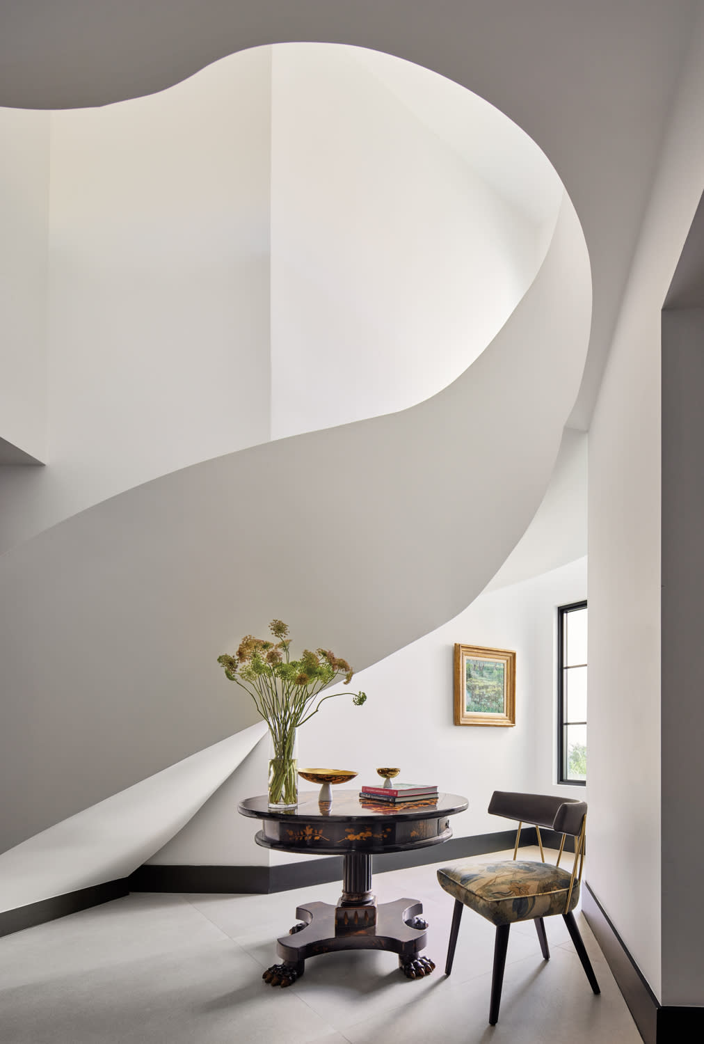 A modern interior features a curved staircase above a dark wood table with a vase of flowers and bowls. A patterned chair and framed artwork complete the scene. Minimalist and elegant.