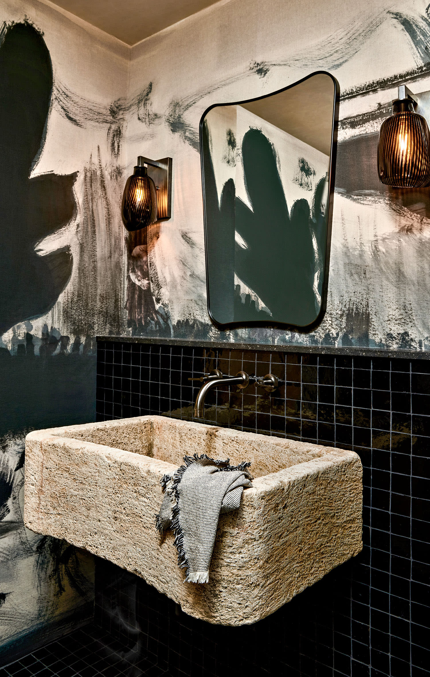 Rustic bathroom with a rough stone sink, black tiled backsplash, and abstract wall art. Warm light from artistic sconces creates an inviting ambiance.