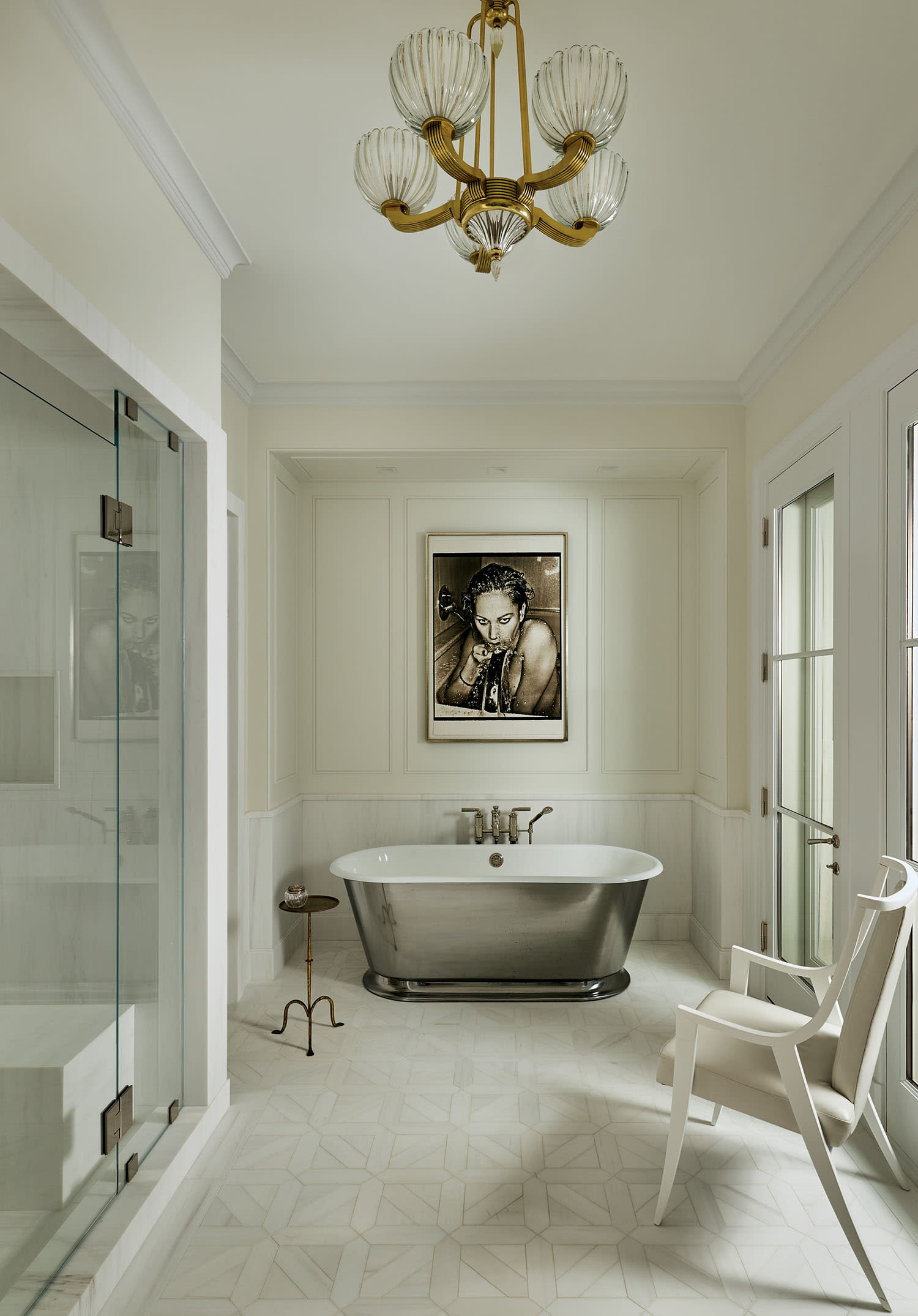 Elegant bathroom with a central freestanding bathtub, artistic wall portrait, and luxurious chandelier. Neutral tones create a serene ambiance.