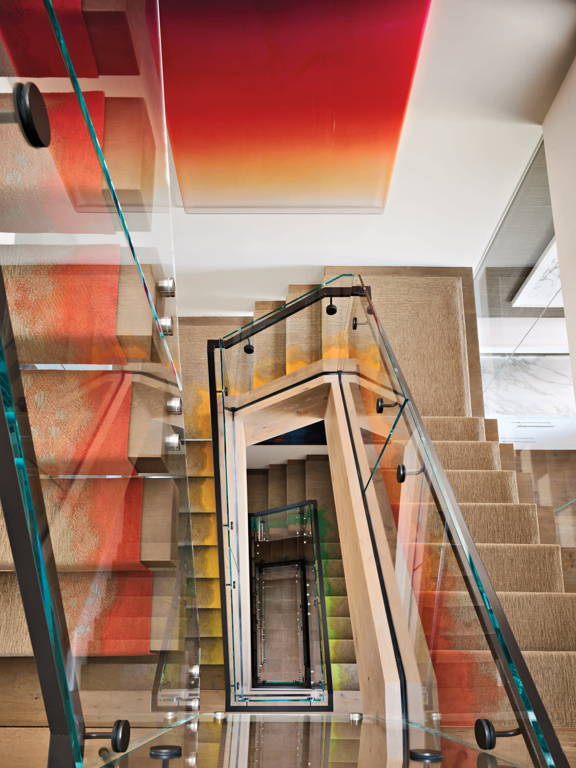 A spiral staircase with light wood steps and glass railings viewed from above. Colorful art with red and orange hues adorns the wall, creating a modern feel.