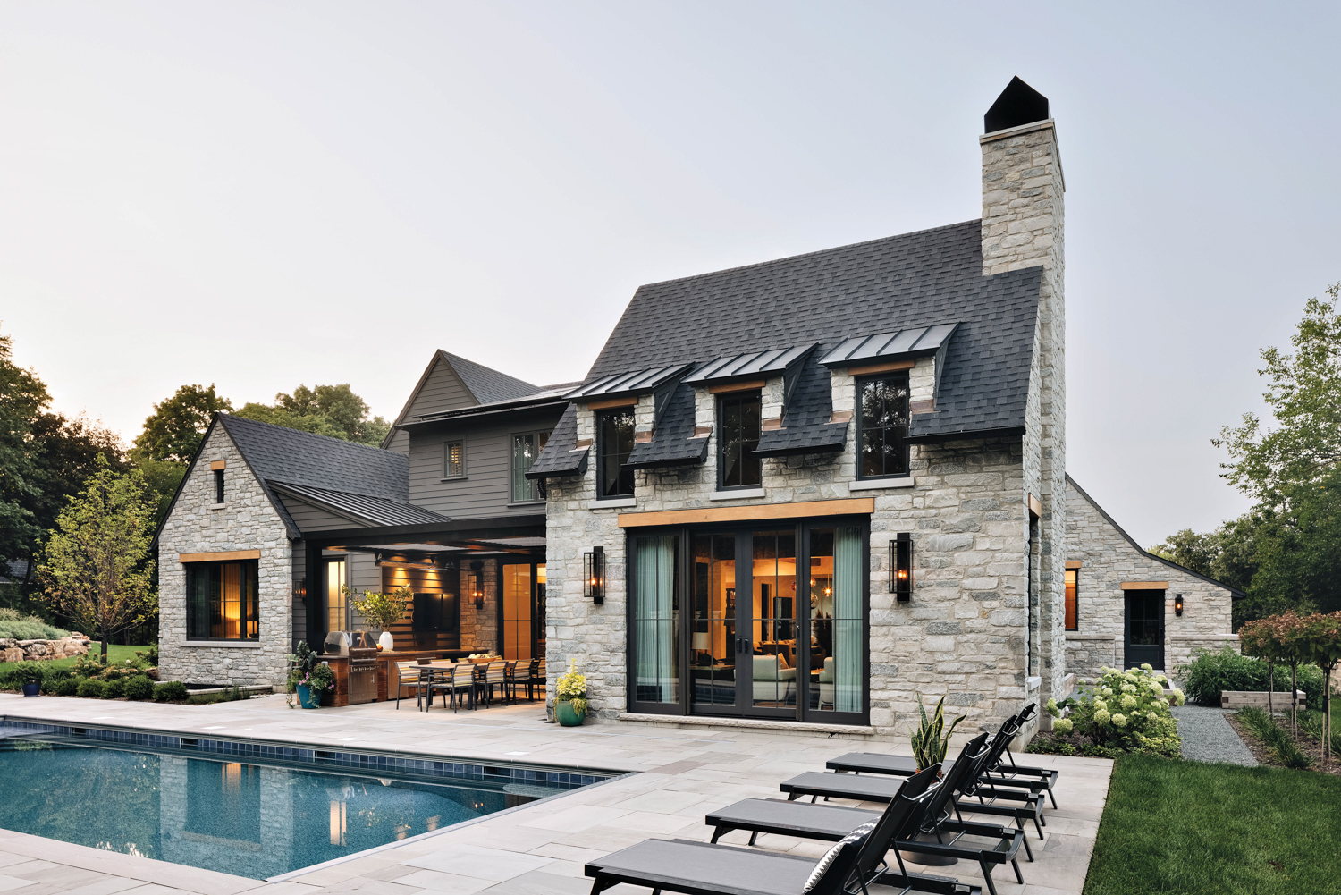 The backyard of a stone home hosts sun loungers and a rectangular pool by Amy Storm