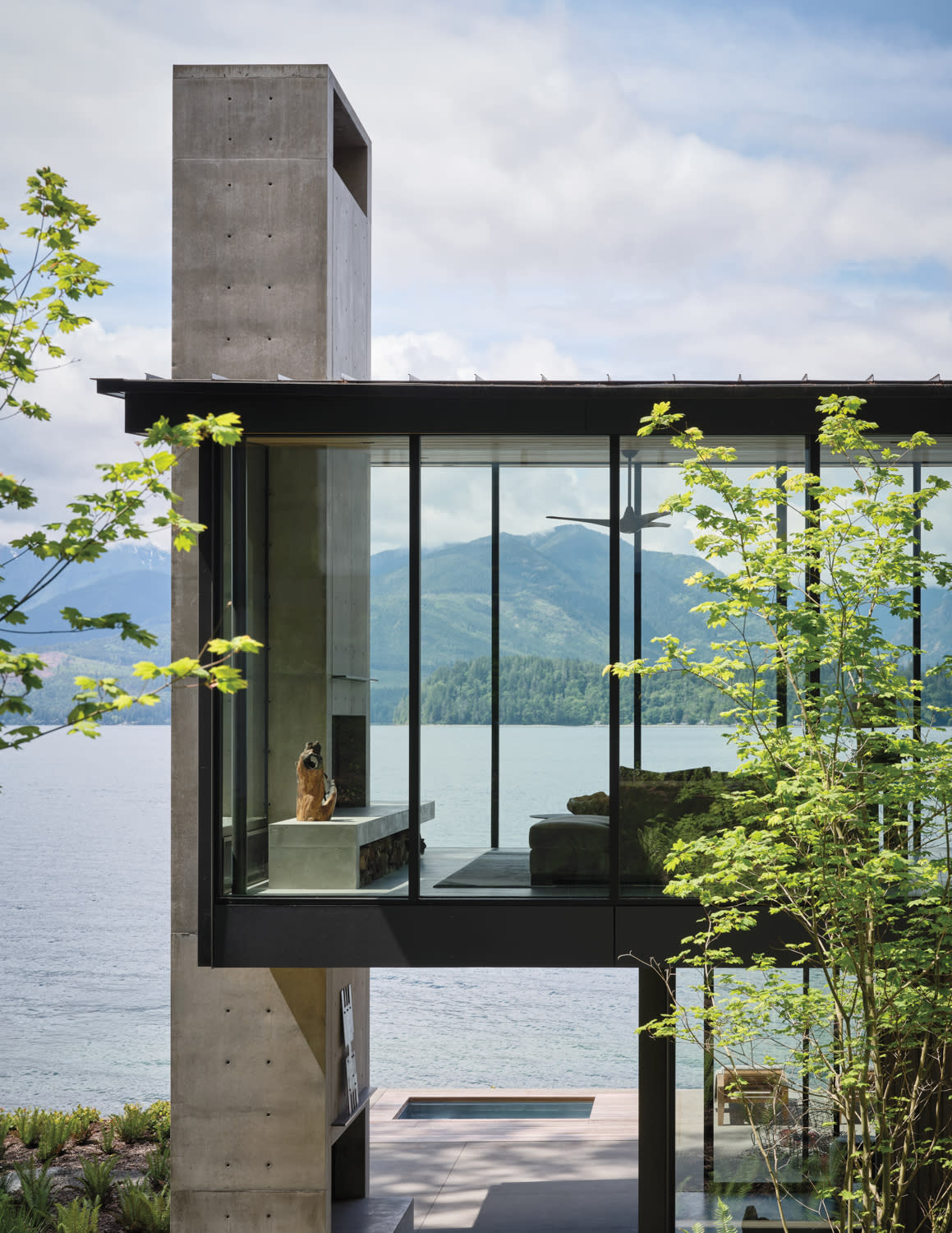 Modern glass house by a serene lake, with mountains in the background. A dog sits inside near a concrete wall, surrounded by lush green trees.