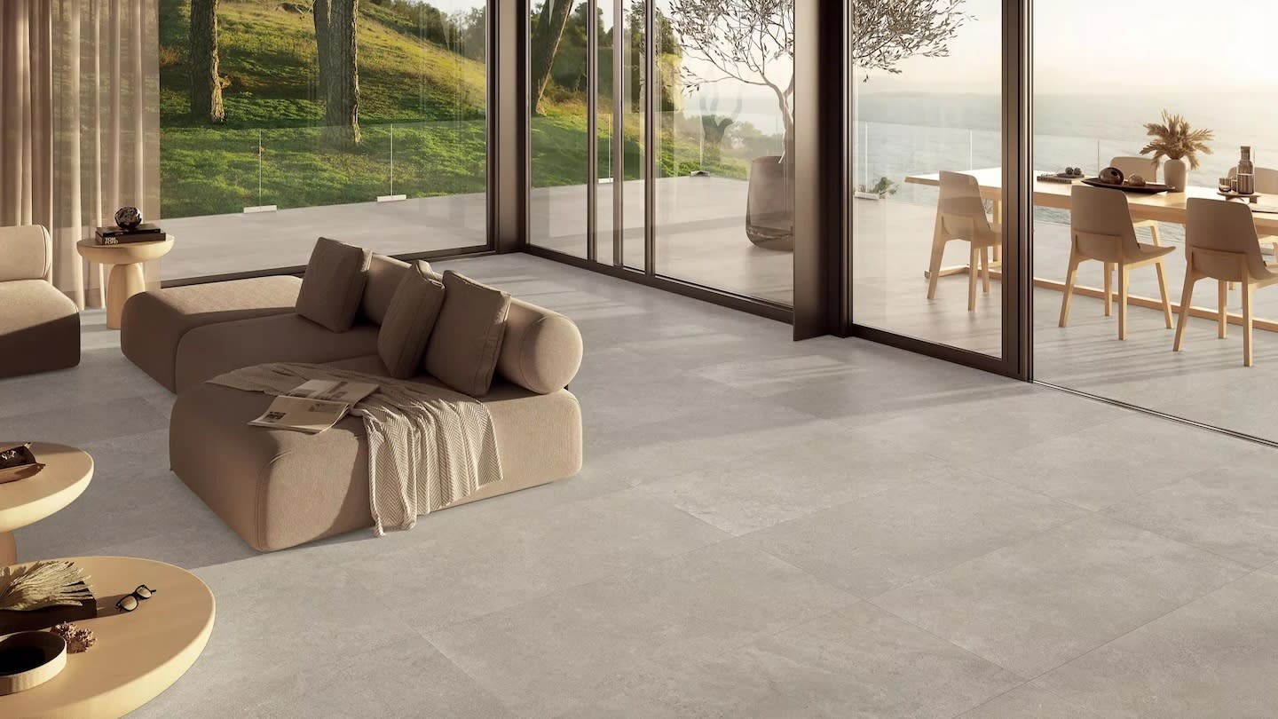 Modern living room with neutral tones, large windows showcasing a serene outdoor view of grass and water. Features a cozy sofa, wooden tables, and elegant decor.