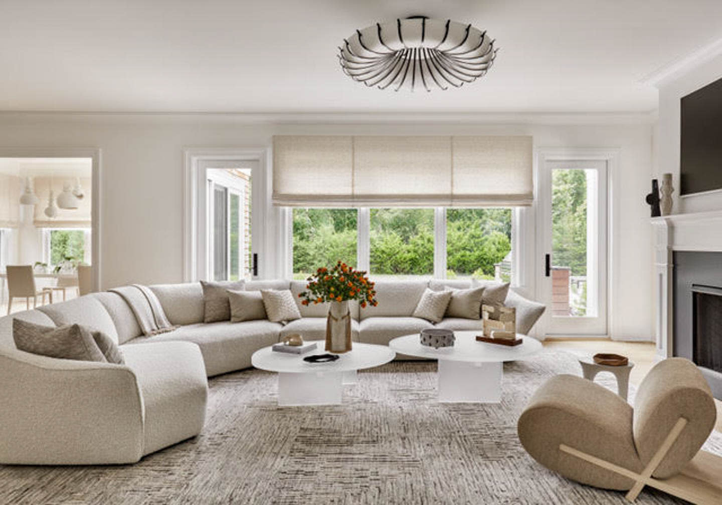 Modern living room with cream tones, featuring a curved sofa, central coffee tables with decor, soft lighting, and large windows overlooking greenery.