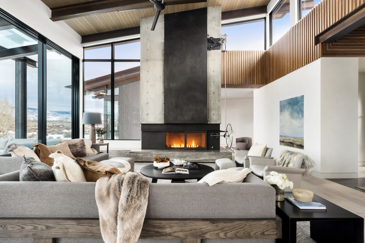 Sophisticated living room with modern grey sofas, a central round table, and a tall fireplace. Floor-to-ceiling windows showcase a scenic winter view.