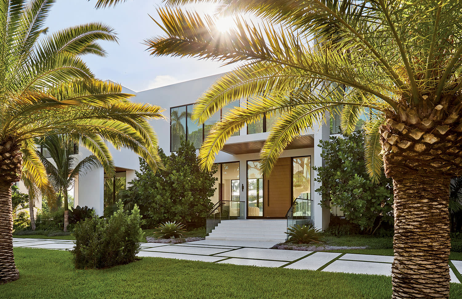 Modern white house framed by lush palm trees and a sunlit garden. The entrance features large windows and a welcoming walkway, conveying a serene, tropical feel.