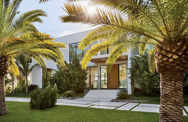 Modern white house framed by lush palm trees and a sunlit garden. The entrance features large windows and a welcoming walkway, conveying a serene, tropical feel.