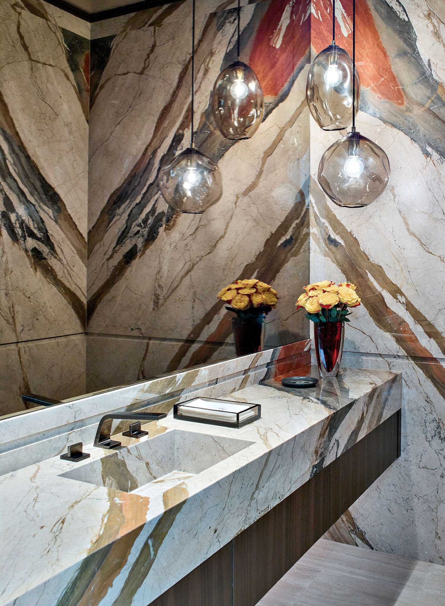 Luxurious bathroom with marble surfaces, abstract patterns, and a sleek black toilet. Three pendant lights, yellow flowers in a vase, modern yet warm.