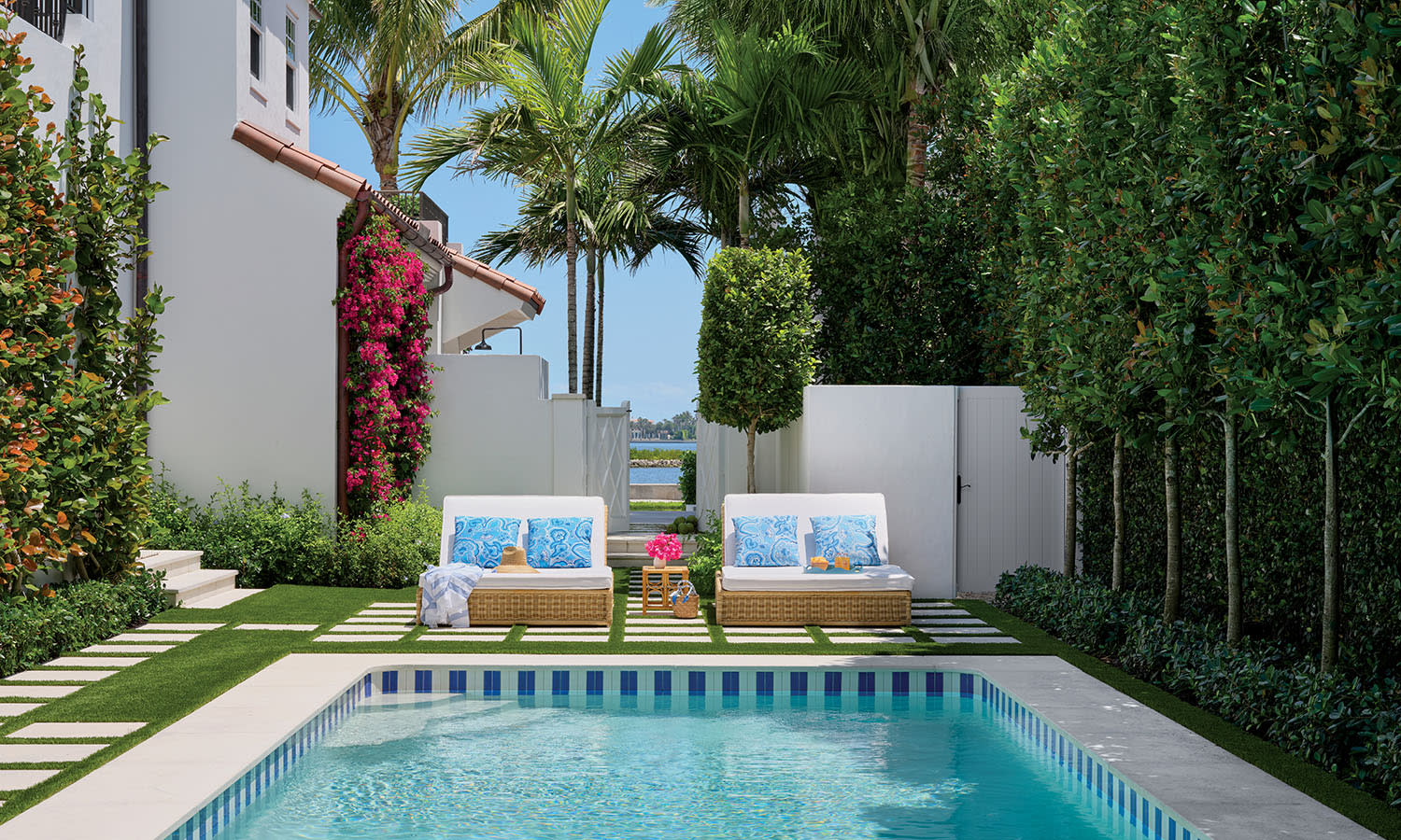 A serene outdoor pool setting with palm trees and a blue sky. Two cushioned lounge chairs with colorful pillows face the pool, exuding a relaxing vibe.