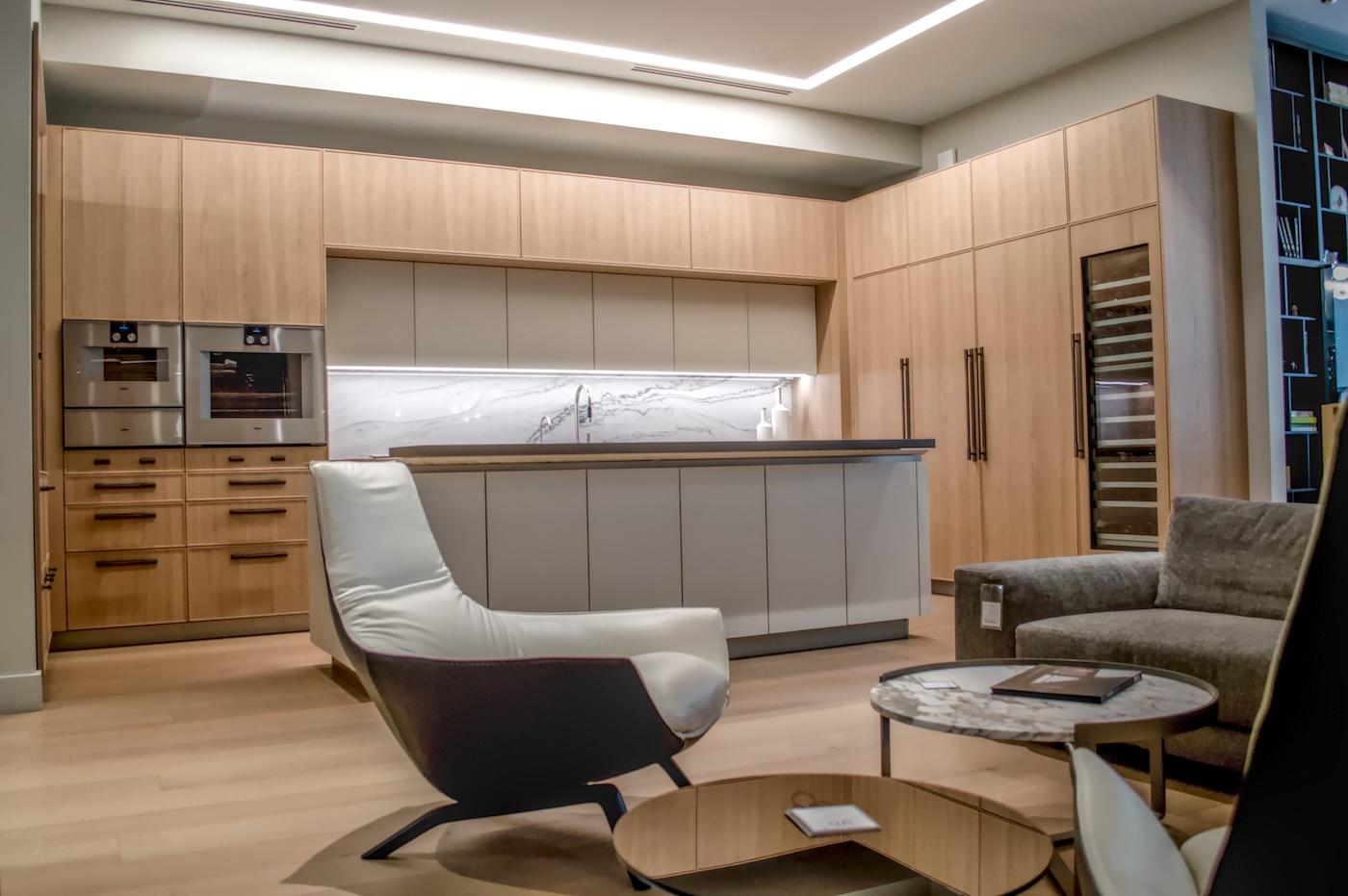 Modern kitchen with light wood cabinets, marble backsplash, and sleek appliances. A cozy seating area features a white armchair and gray sofa.