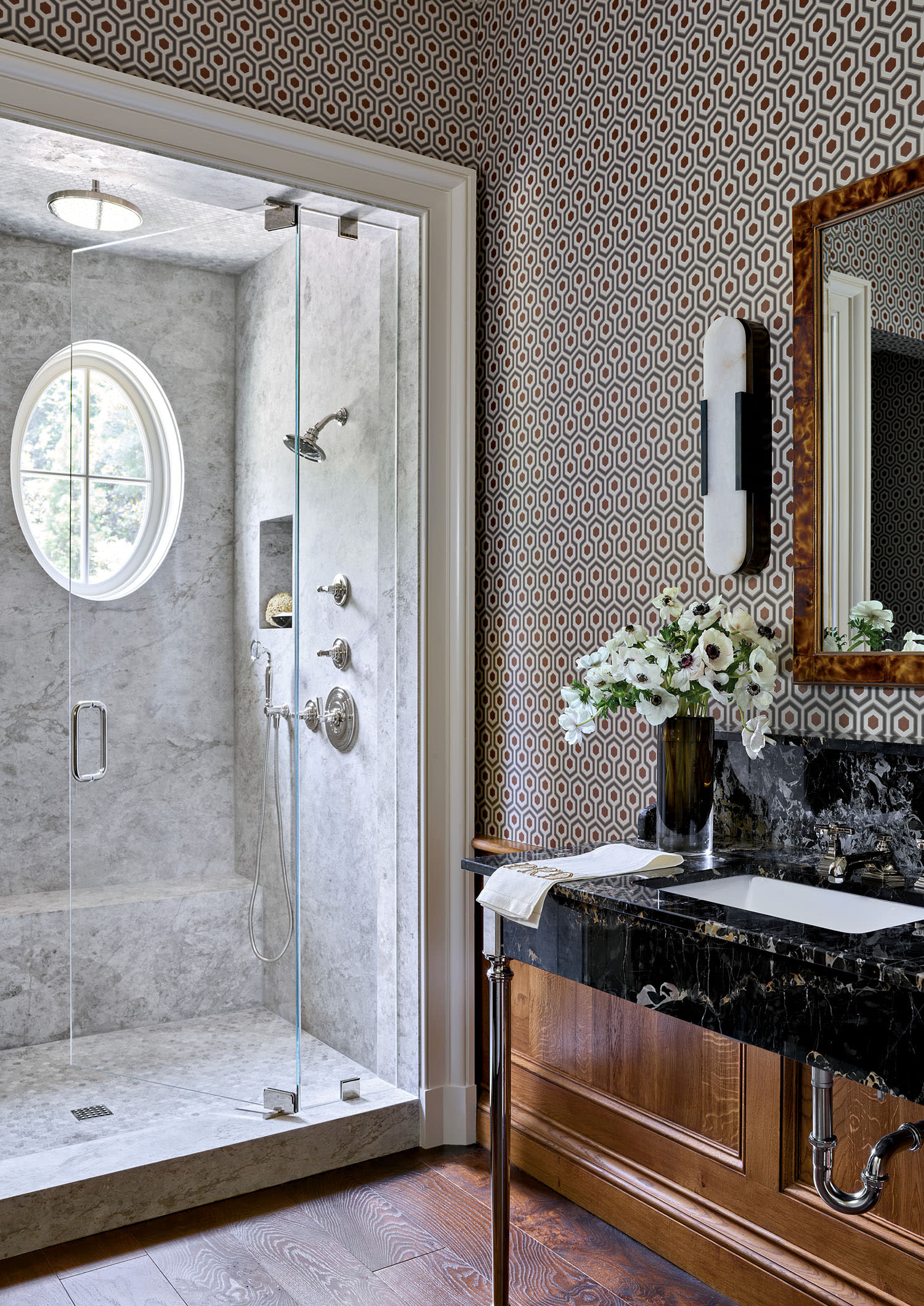 Elegant bathroom featuring a glass-door marble shower, oval window, patterned wallpaper, dark countertop with wood vanity, and a vase of white flowers.