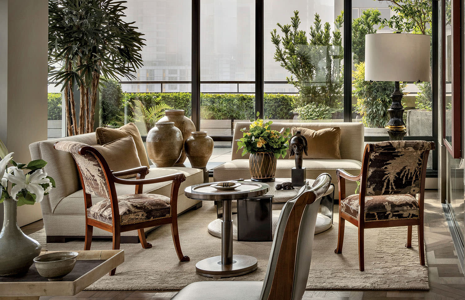Luxurious living room with large windows, beige sofas, patterned armchairs, potted plants and a lamp