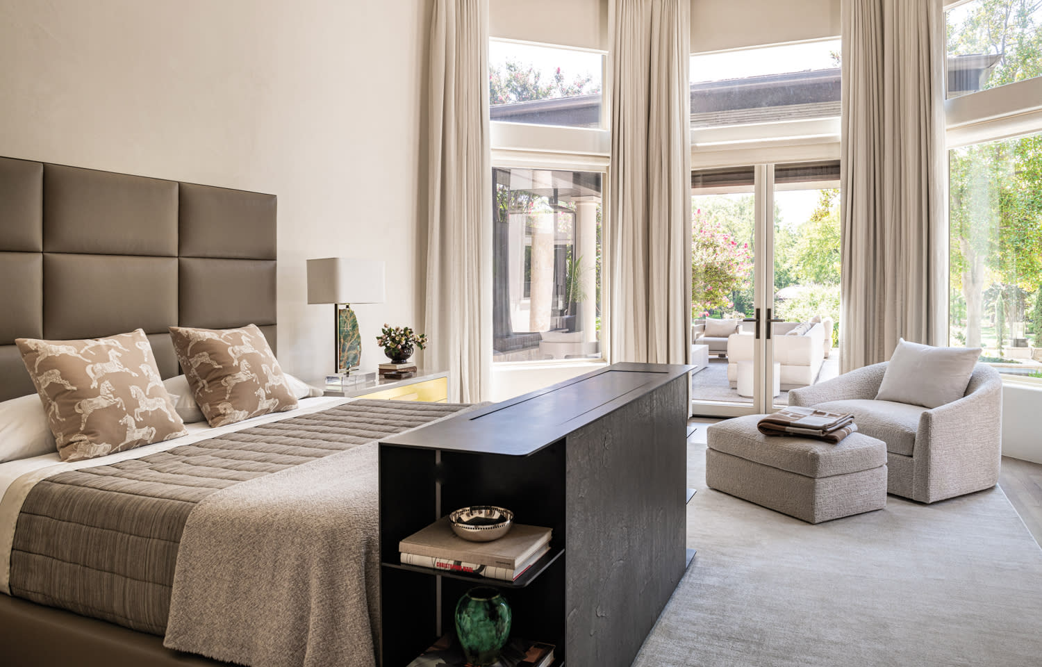 Luxurious bedroom with a large padded headboard, neutral tones, and animal-patterned pillows. Tall windows, cozy chaise lounge, and a peaceful, airy ambiance.