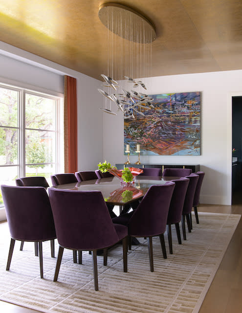 A modern dining room with a table surrounded by purple velvet chairs, a contemporary chandelier, a large painting, and floor-to-ceiling windows with red curtains.