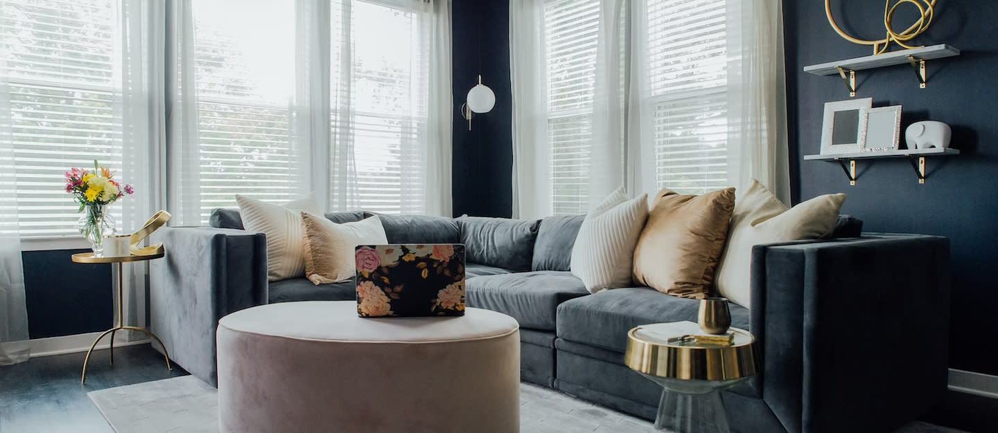 Stylish living room with dark walls, a gray sectional sofa adorned with soft pillows, a floral laptop on an ottoman, gold accent tables, and vibrant flowers.