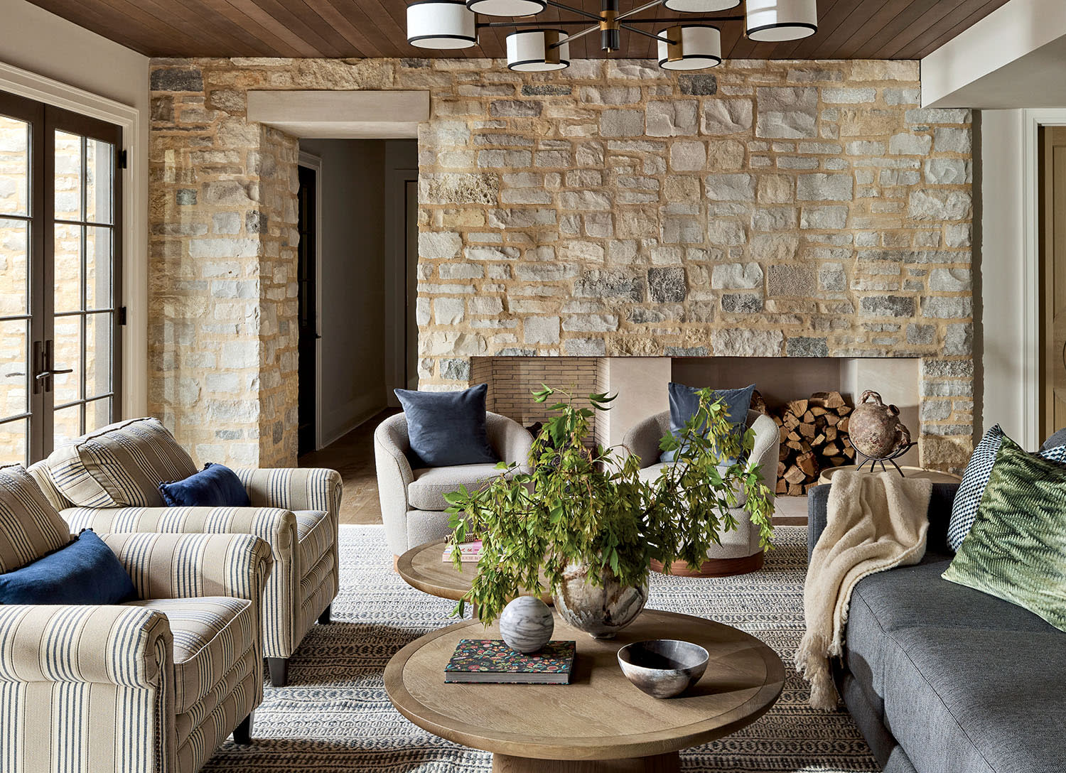 Warm living room with stone wall, modern chandelier, and cozy furniture. Striped and gray sofas, wood tables, plants, and log-filled fireplace create a welcoming feel.
