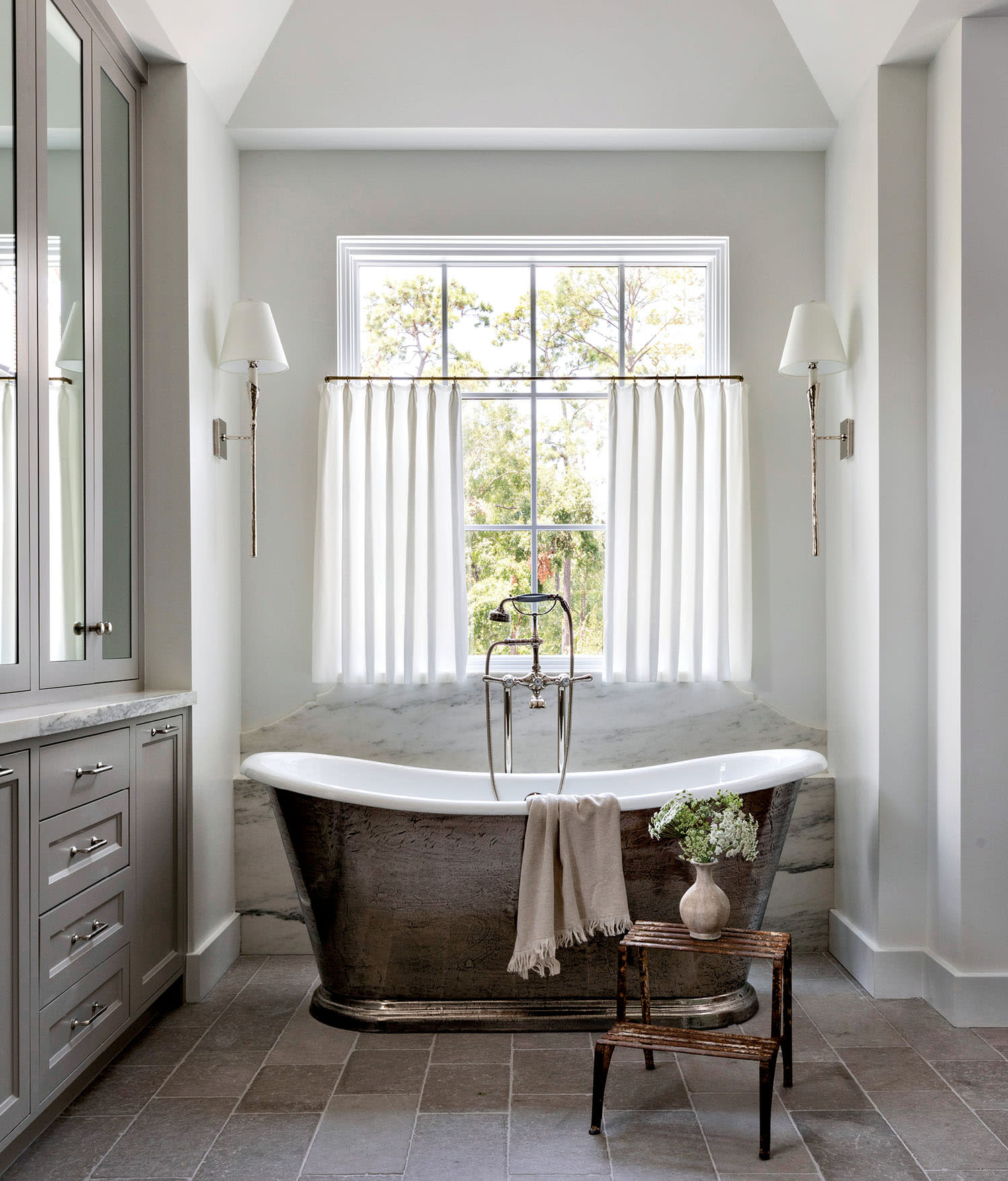 Elegant bathroom with freestanding metallic bathtub under a large window with sheer curtains, flanked by wall sconces; neutral tones evoke tranquility.
