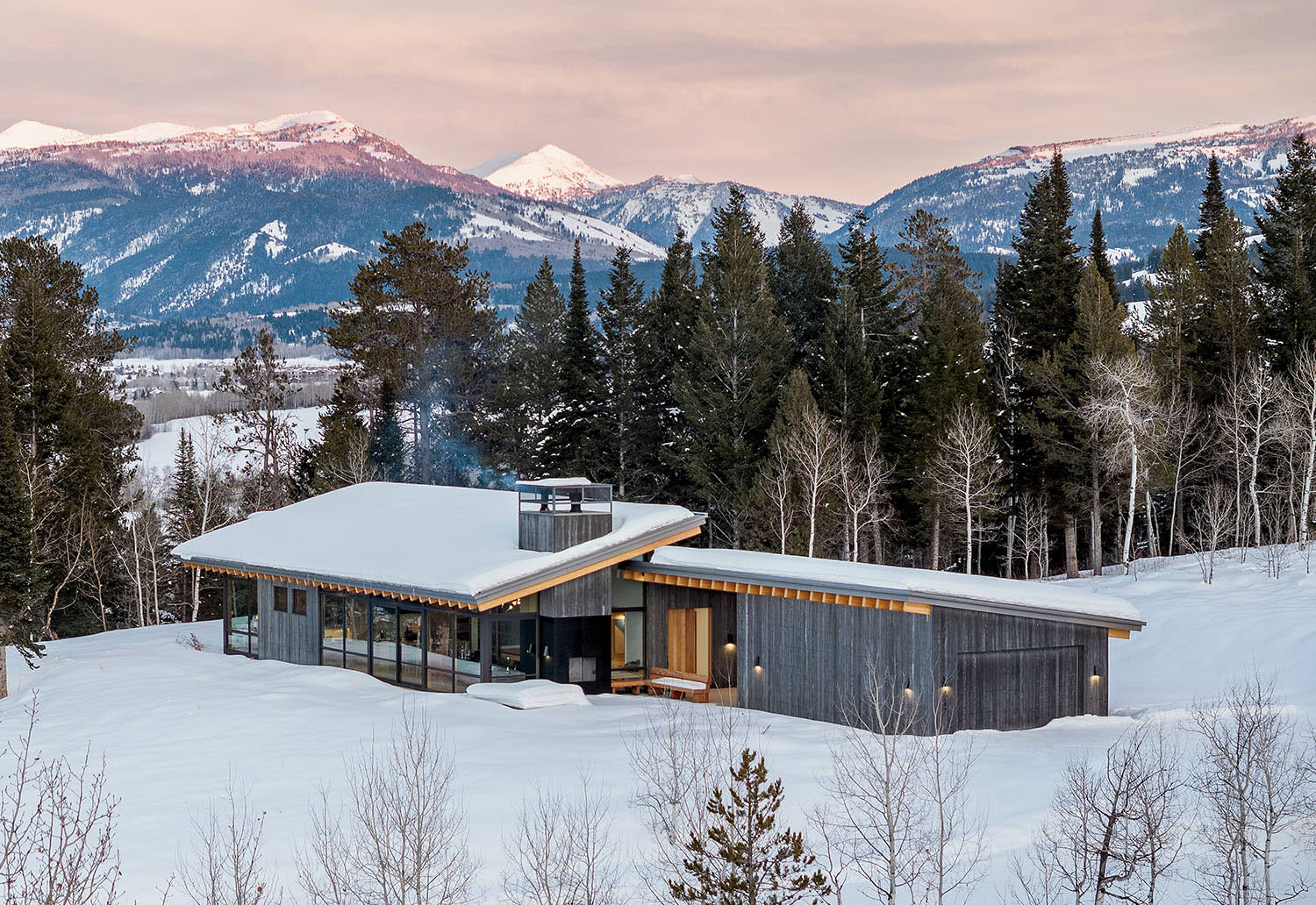 The exterior of a home photographed from uphill, surrounded by snow, trees and mountains.