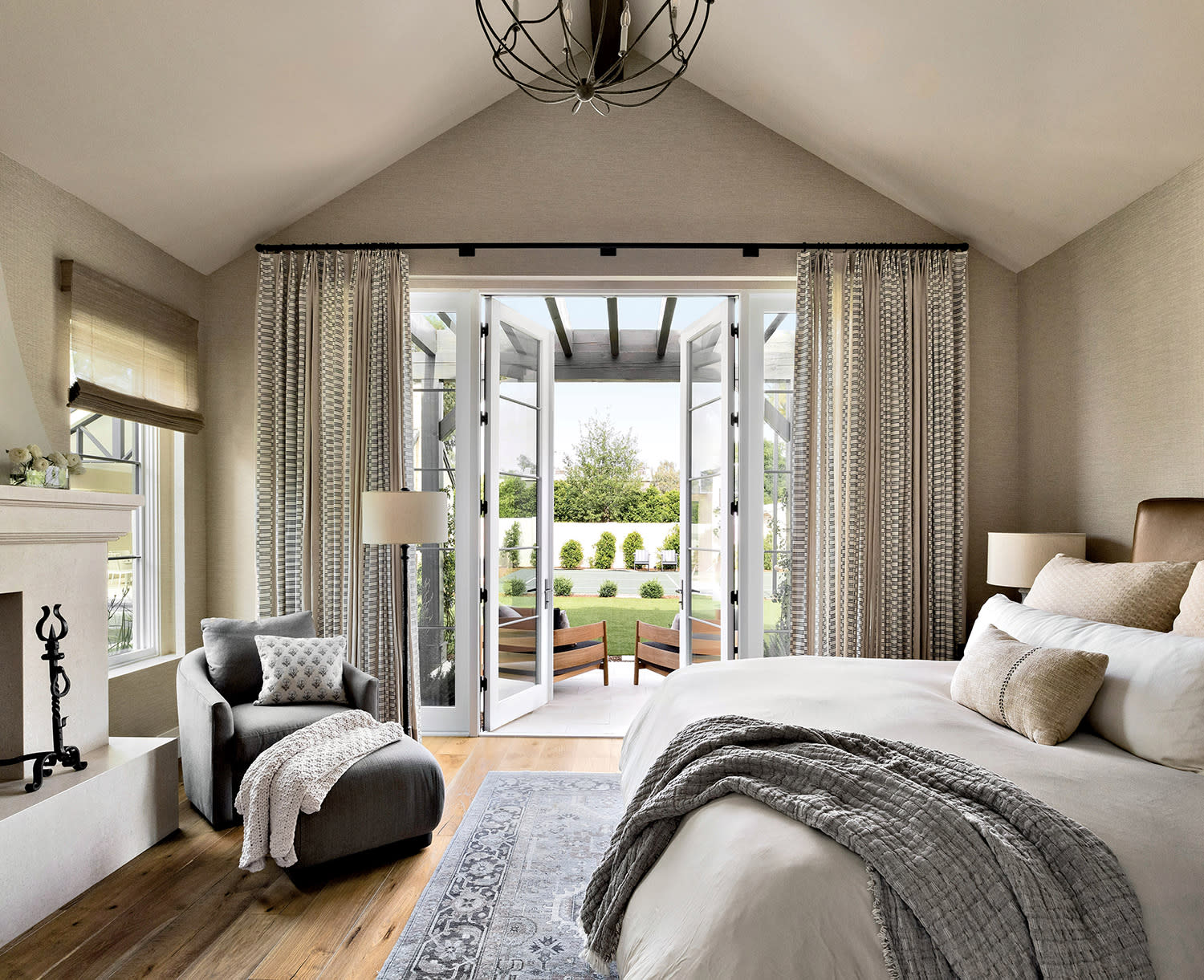 A cozy bedroom with neutral tones, featuring a plush bed, elegant chandelier, armchair by a fireplace, and French doors opening to a lush garden view.