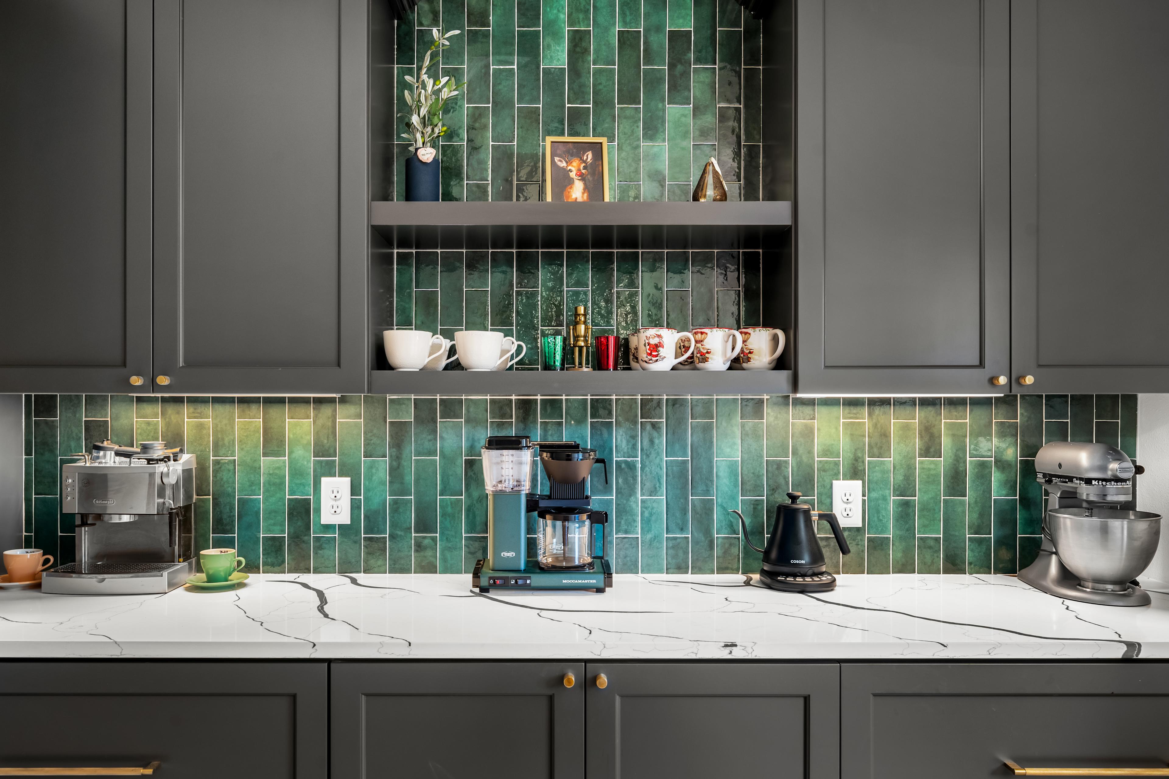 This stylish coffee station features deep green tile backsplash,
dark cabinetry with gold accents, and elegant open shelving. A sleek marble countertop provides
ample space for coffee makers and decor, blending functionality with modern sophistication.