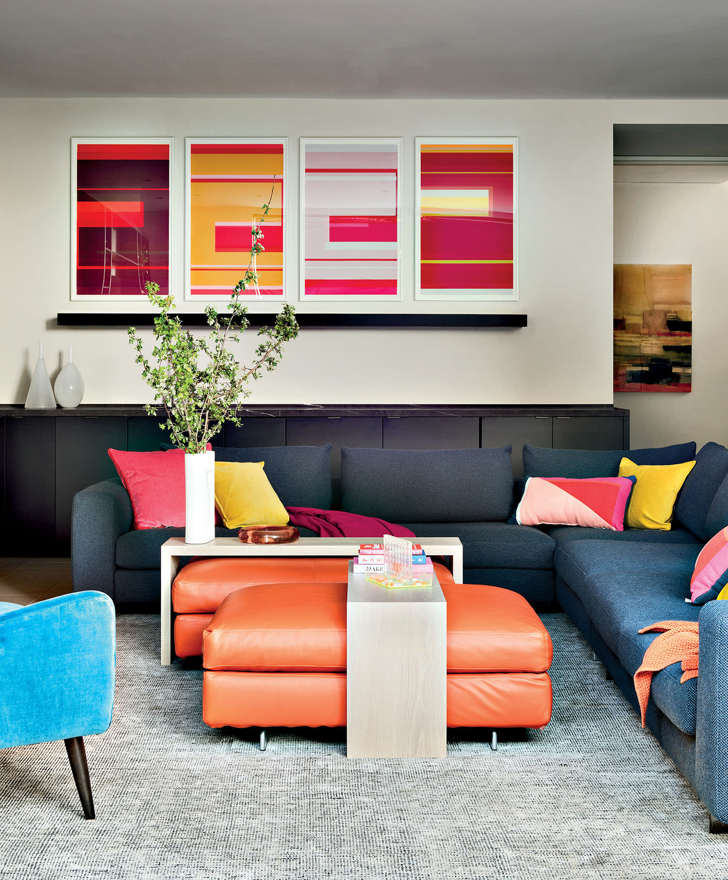 Modern living room with a blue sectional sofa, colorful pillows, and an orange ottoman. Vibrant artwork adorns the walls, creating a lively ambiance.