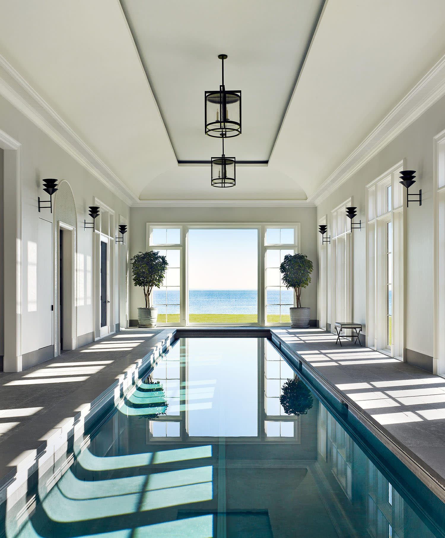 Luxurious indoor pool with tall windows, offering a stunning ocean view. Symmetrical design features potted plants and ceiling lights, creating a serene ambiance.