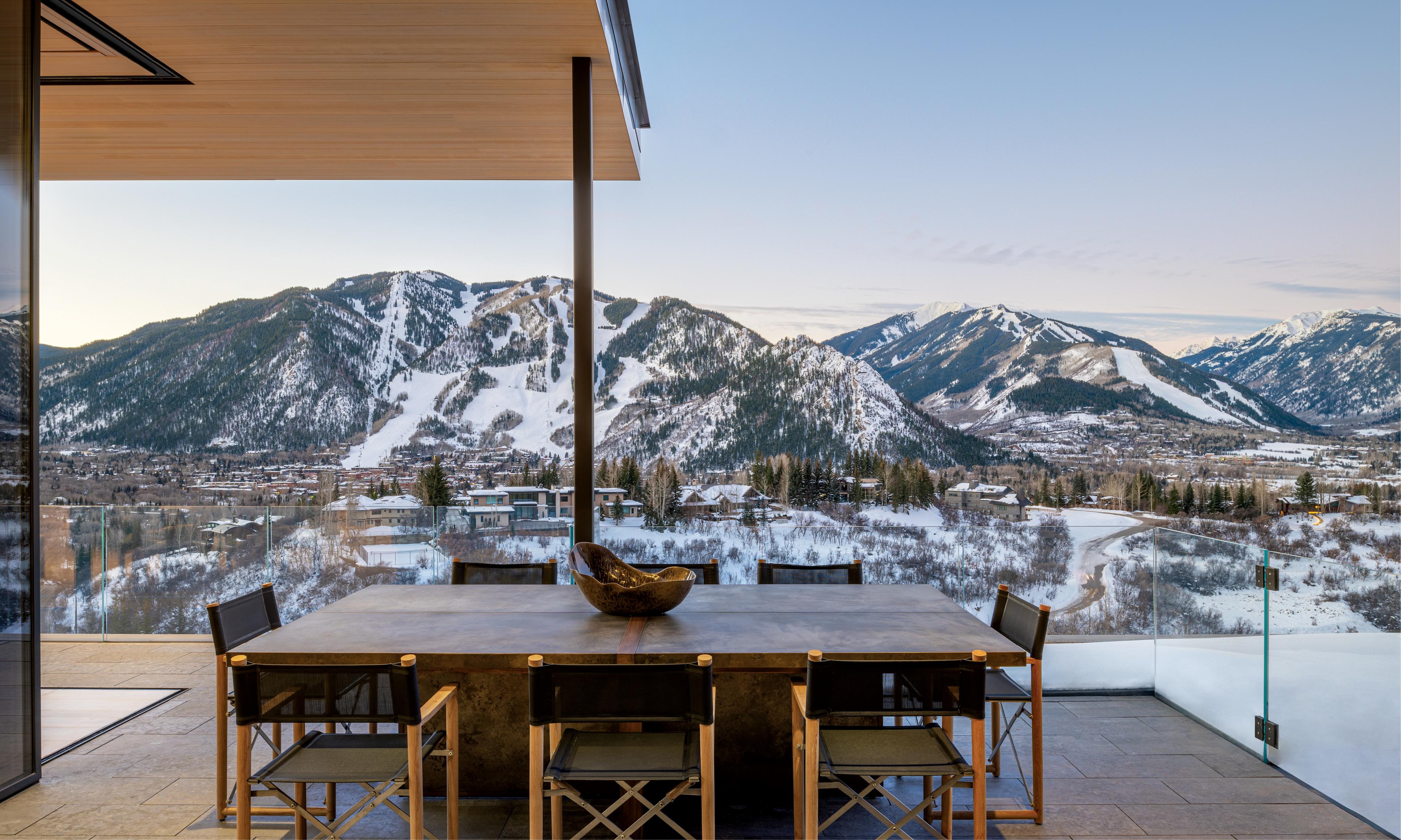 A modern patio with a wooden table and chairs overlooks a stunning snowy mountain landscape. The scene is tranquil, with clear skies and soft lighting.