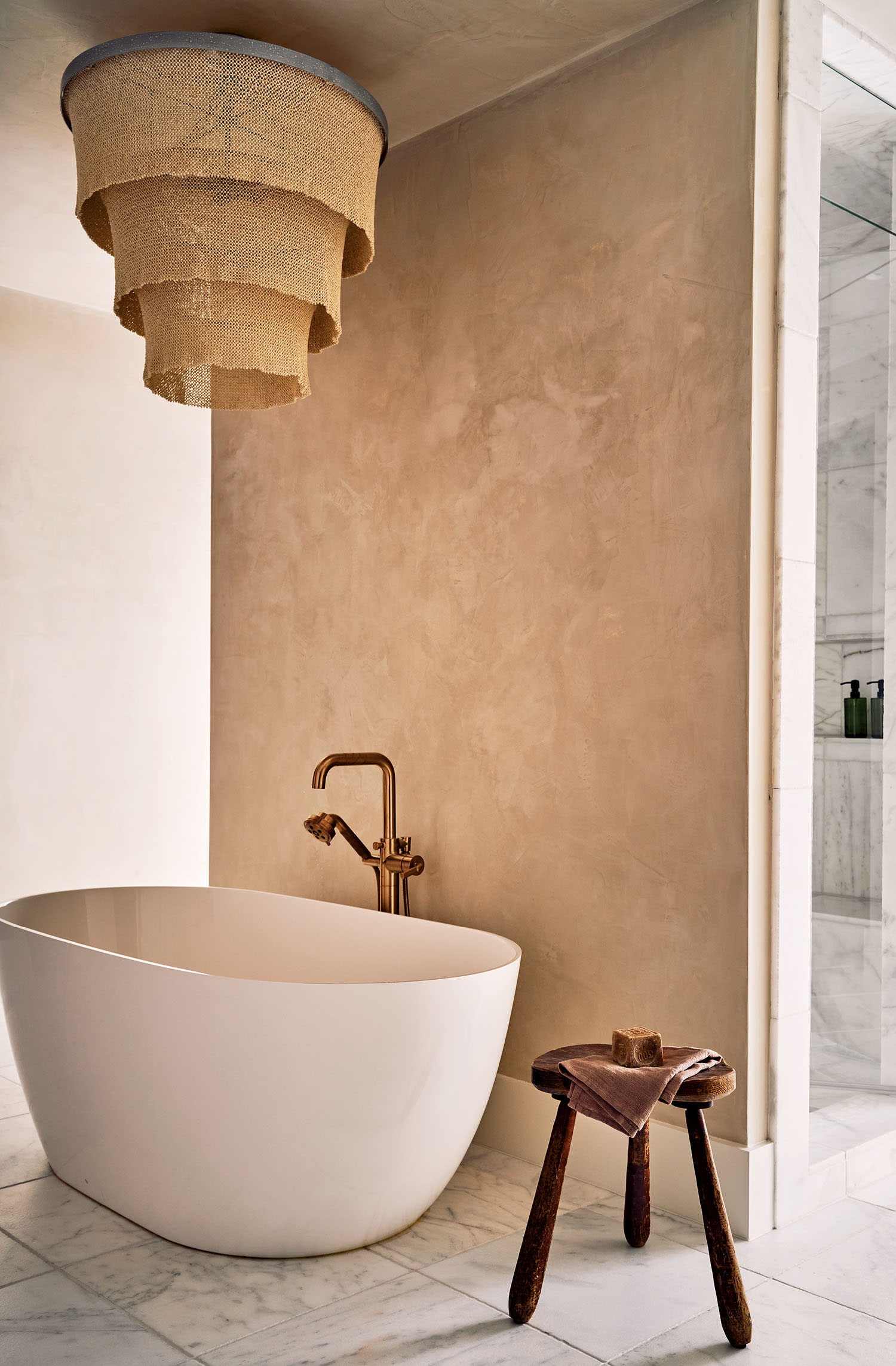 A minimalist bathroom with a white oval tub and copper faucet. A rustic stool holds folded towels. A layered beige chandelier adds warmth above.