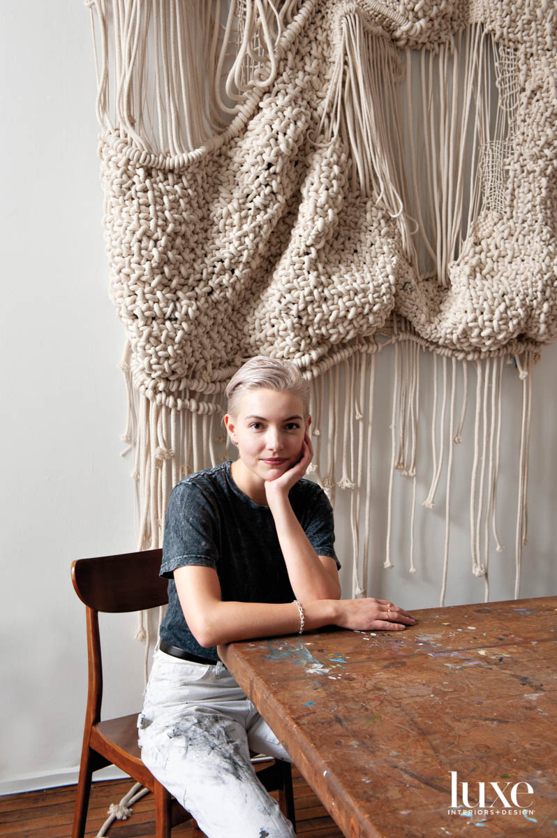 A woman with short hair since at a table in front of a rope hanging.