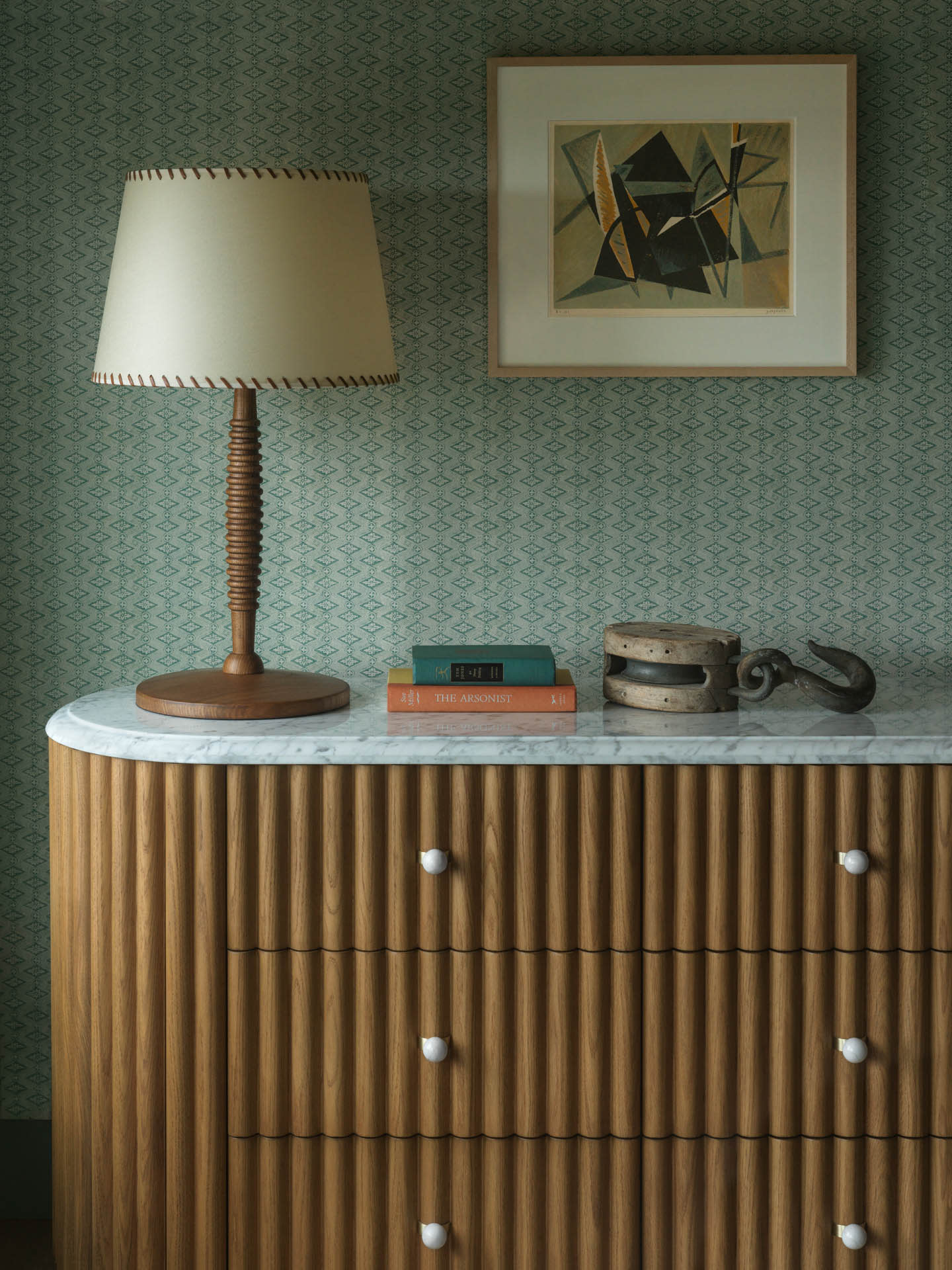 A wooden dresser with a marble top features a table lamp, stacked books, and a vintage pulley. A modern abstract painting adorns the patterned wall.
