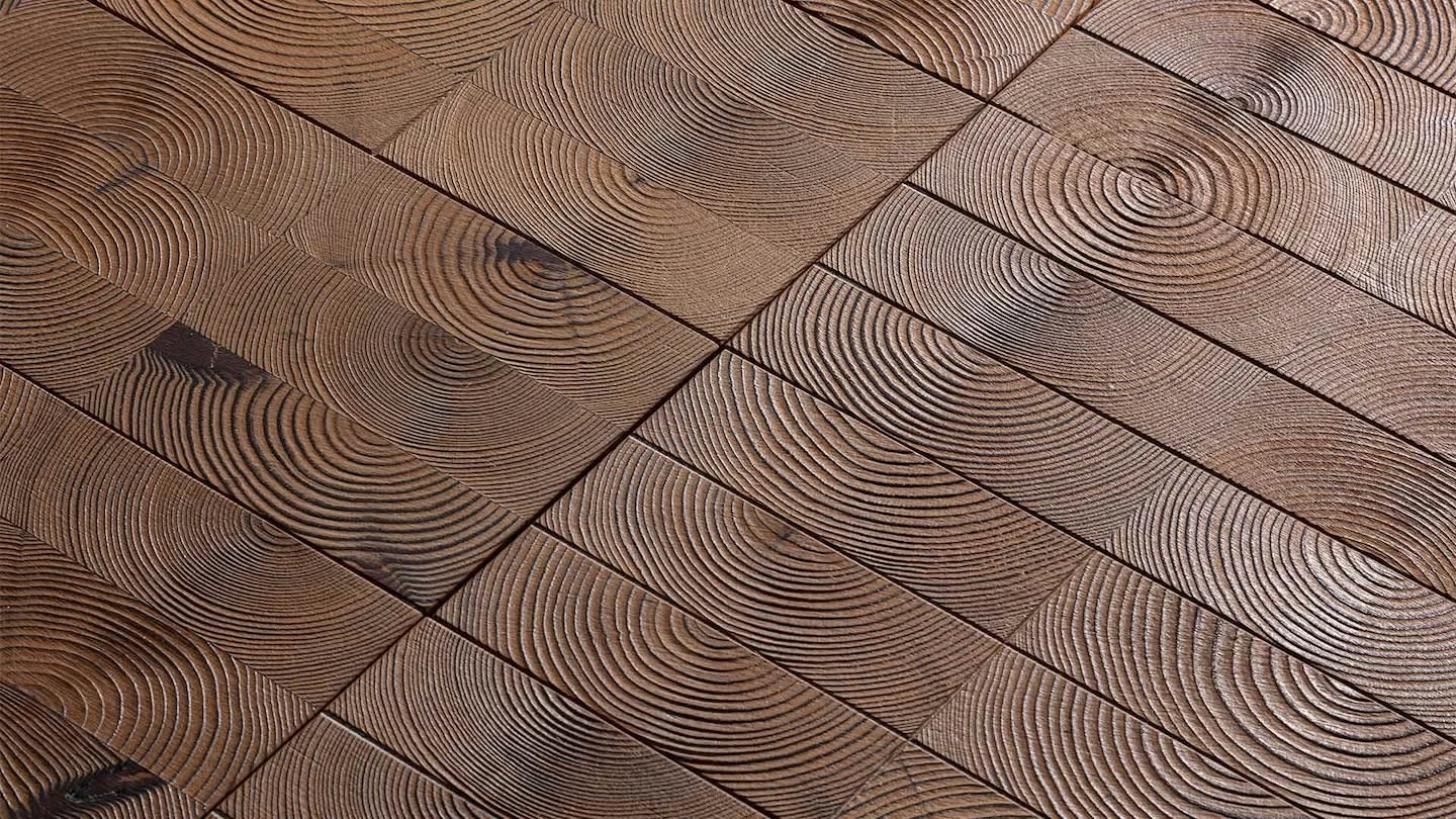 Close-up of a wooden floor with a geometric pattern of square tiles. Each tile features distinct circular wood grain rings, adding a rustic and textured appearance.