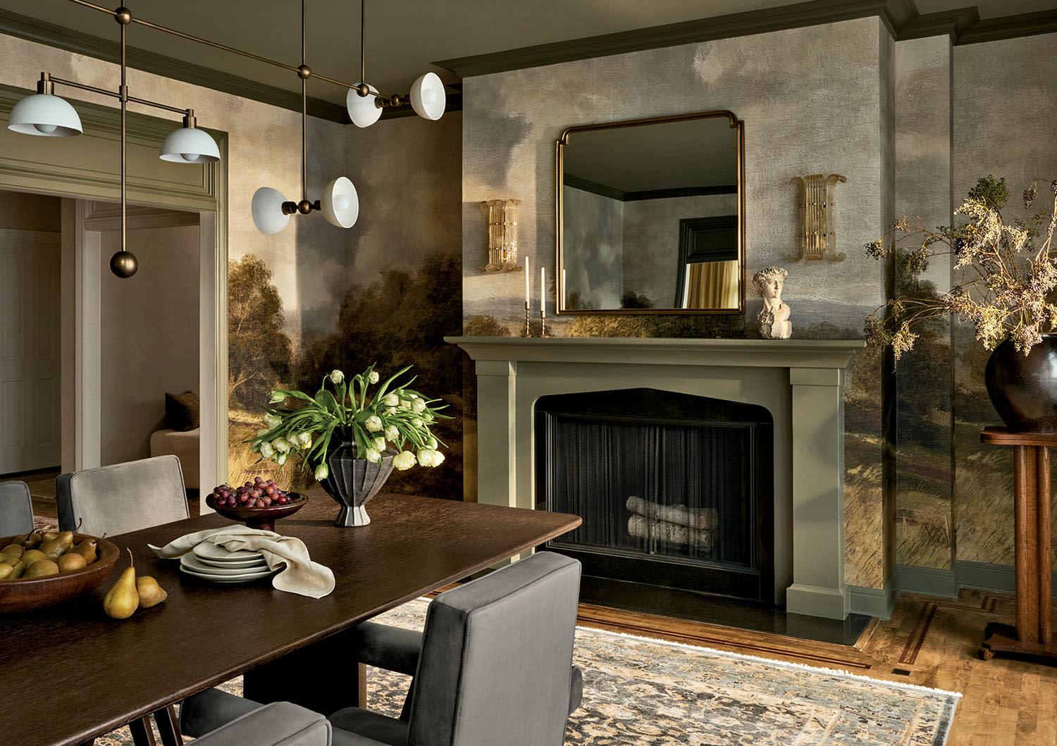 Elegant dining room with a rustic, nature-themed mural. Features a wooden table, gray chairs, ornate fireplace, and decorative lighting, evoking warmth.