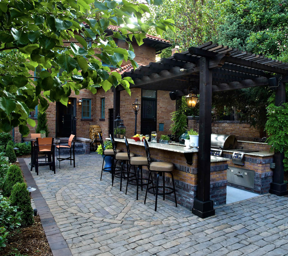 Private brick patio featuring grill and bar, enclosed by lush greenery, pergola overhead.