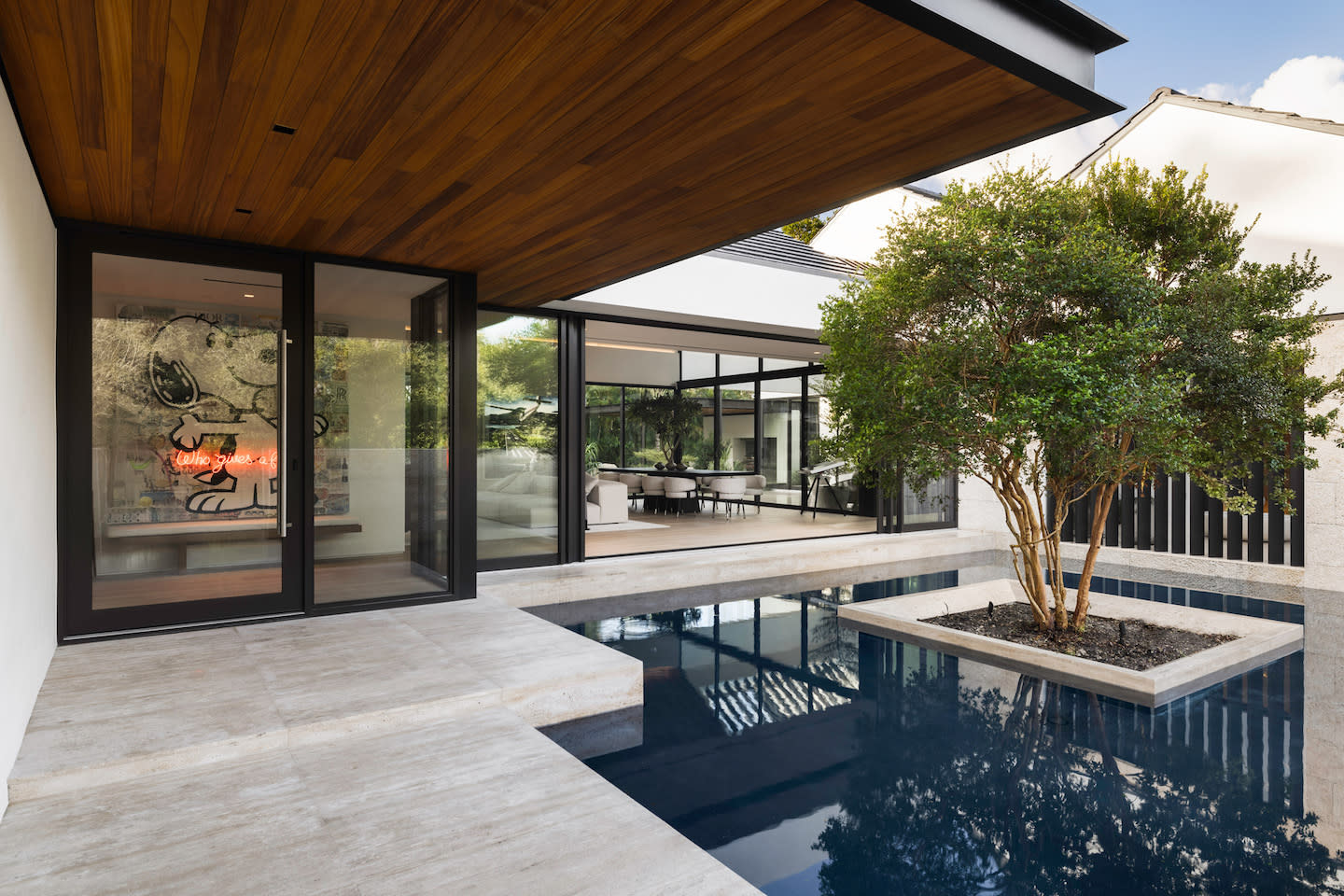 Zen courtyard scene featuring a sleek travertine walkway that hovers over a dark reflecting pool, its still surface cradling an island planter with a leafy tree. The space flows into an open-concept living area through floor-to-ceiling black-framed glass doors, while a warm cedar soffit and a peek of playful neon artwork add cozy character to the minimalist architecture.