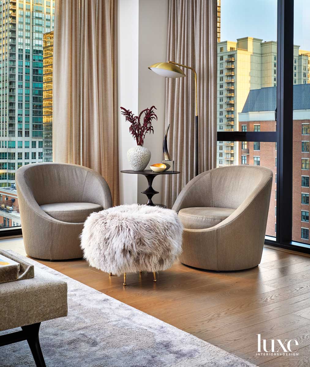 Two swivel chairs with a fur ottoman in between them. There is a circular side table with a floor lamp above it. You see the city through the windows behind the seating arrangement.