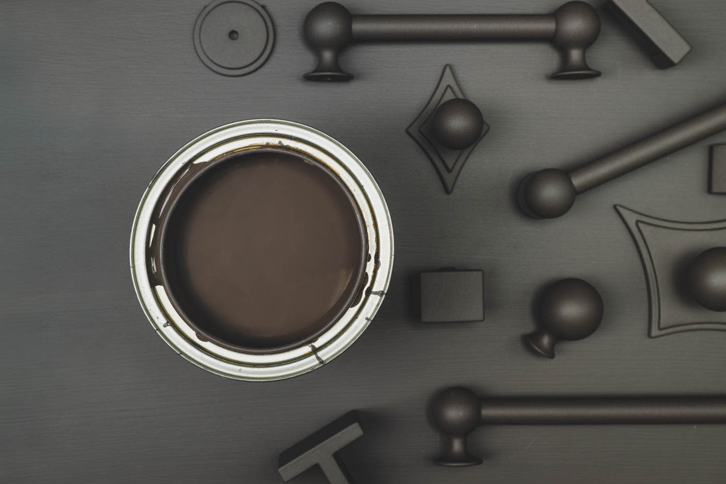Open can of dark paint on a gray surface surrounded by various matte black metal handles and knobs. Industrial, modern aesthetic.