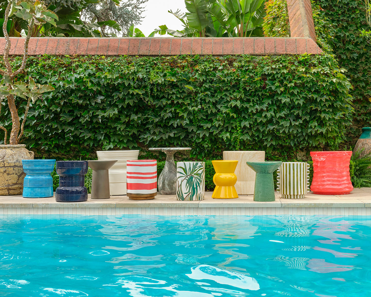 Bright, colorful stools line a poolside with a backdrop of lush, green ivy on a brick wall. The scene conveys a vibrant, summery atmosphere.