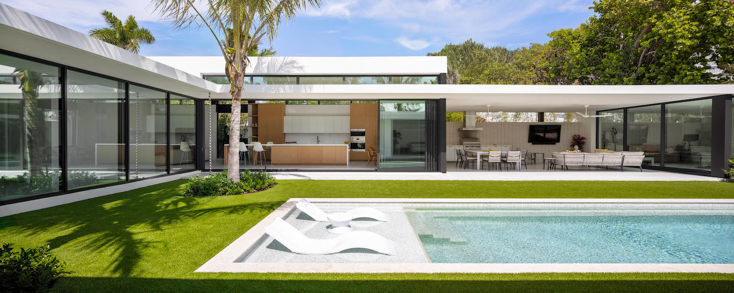 Modern pavilion-style villa with crisp white rooflines and black-framed glass walls surrounding a lush courtyard lawn and rectangular pool, its shallow ledge holding two sculptural in-water loungers. A breezy open kitchen with blond wood cabinetry spills into a shaded alfresco dining and lounge area, complete with ceiling fans and a wall-mounted TV, all framed by palms and leafy greenery beneath a bright blue sky.