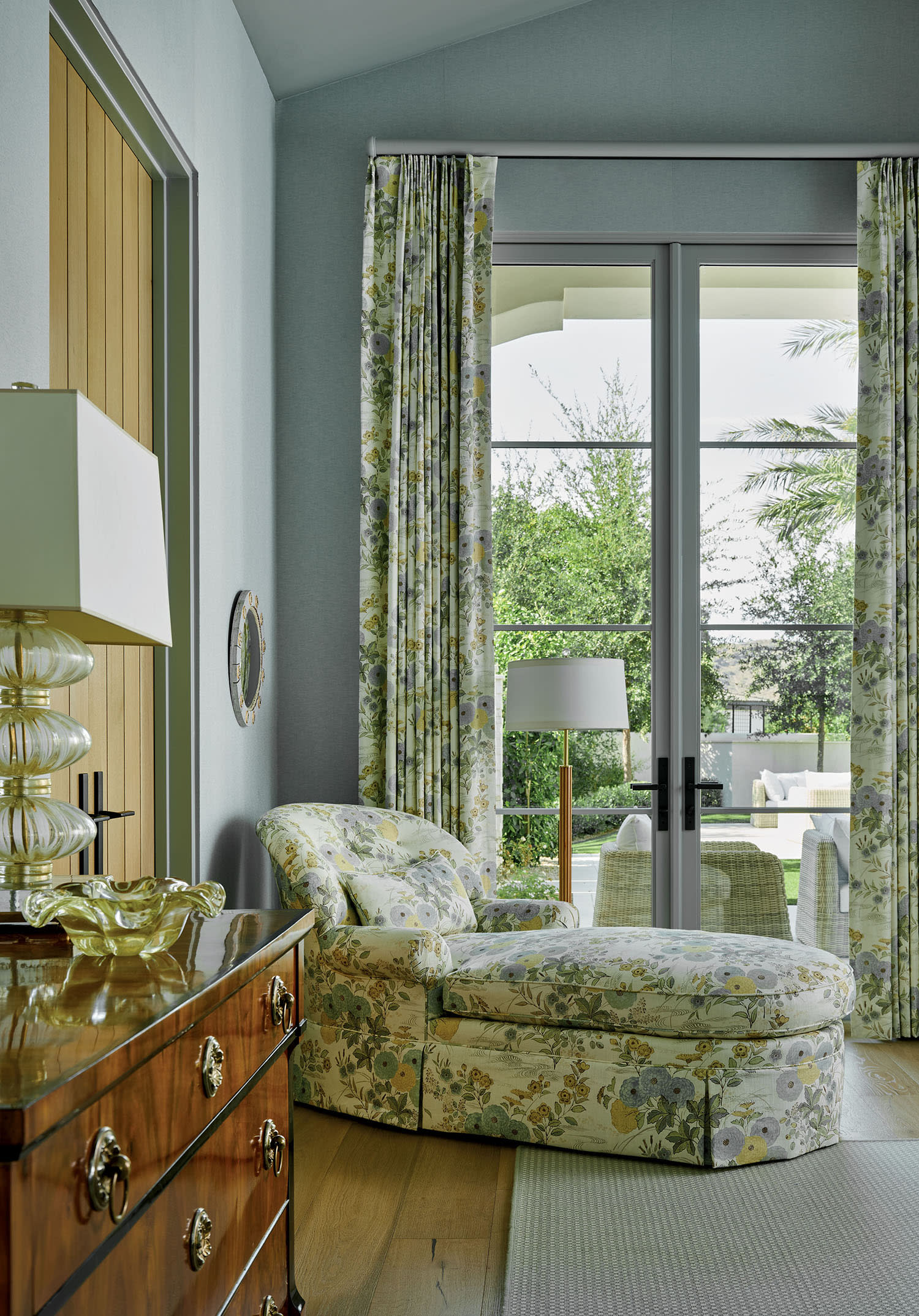 Elegant room with a floral chaise lounge by large windows featuring floral curtains. A wooden dresser with a glass lamp adds warmth and sophistication.