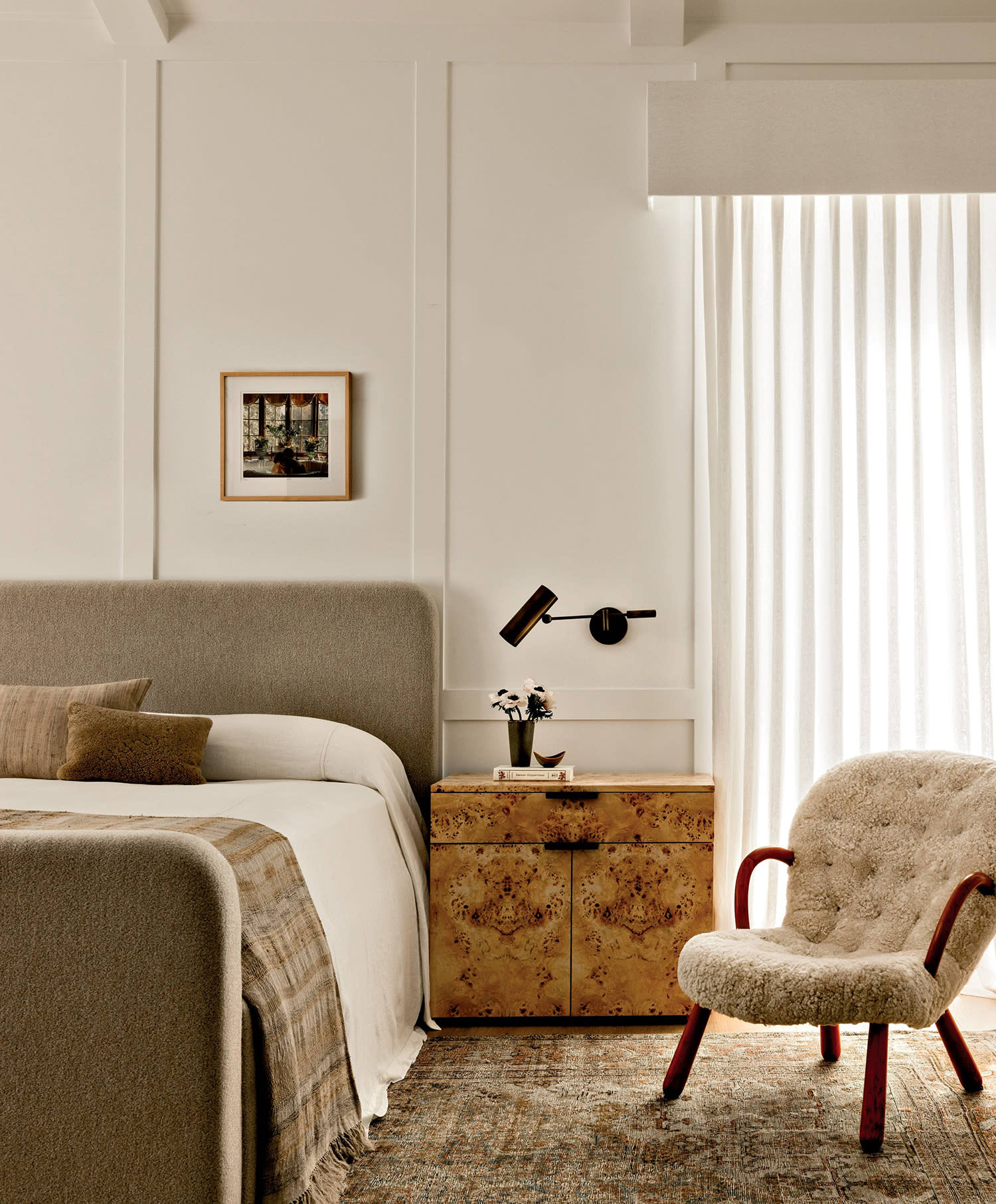 A bed sits against a wall with artwork above the headboard and a wood nightstand and armchair nearby.