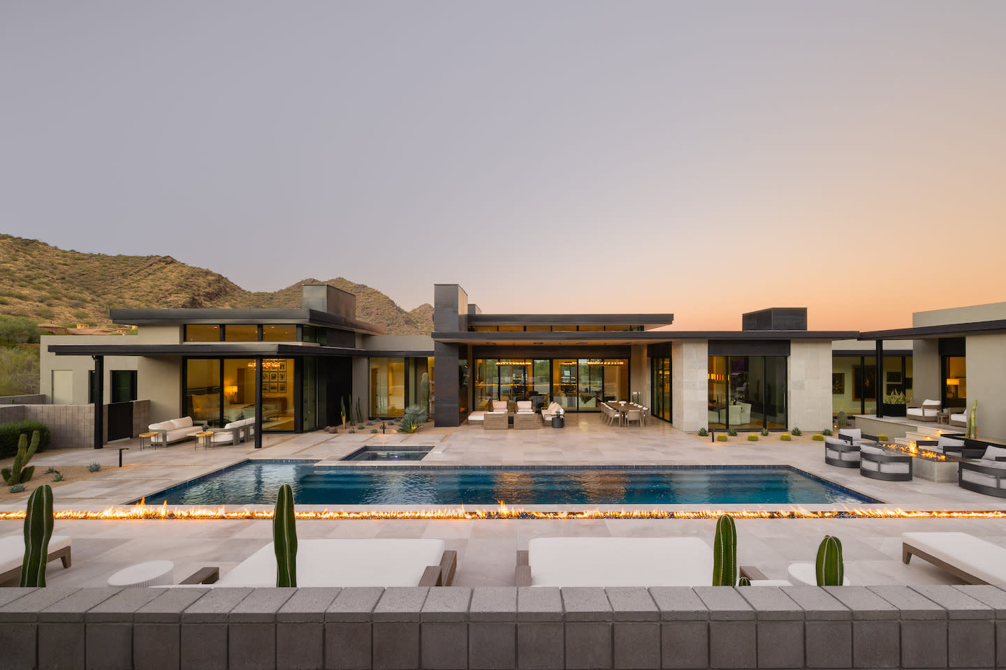 Sleek desert-modern estate at sunset, framed by low mountain ridges in the distance. The scene centers on a long rectangular pool with a continuous ribbon of fire along its edge, flanked by white sun loungers and tall columnar cacti. Floor-to-ceiling glass walls reveal a stylish open-plan living area, while expansive stone decking and cozy seating zones create a resort-like vibe.
