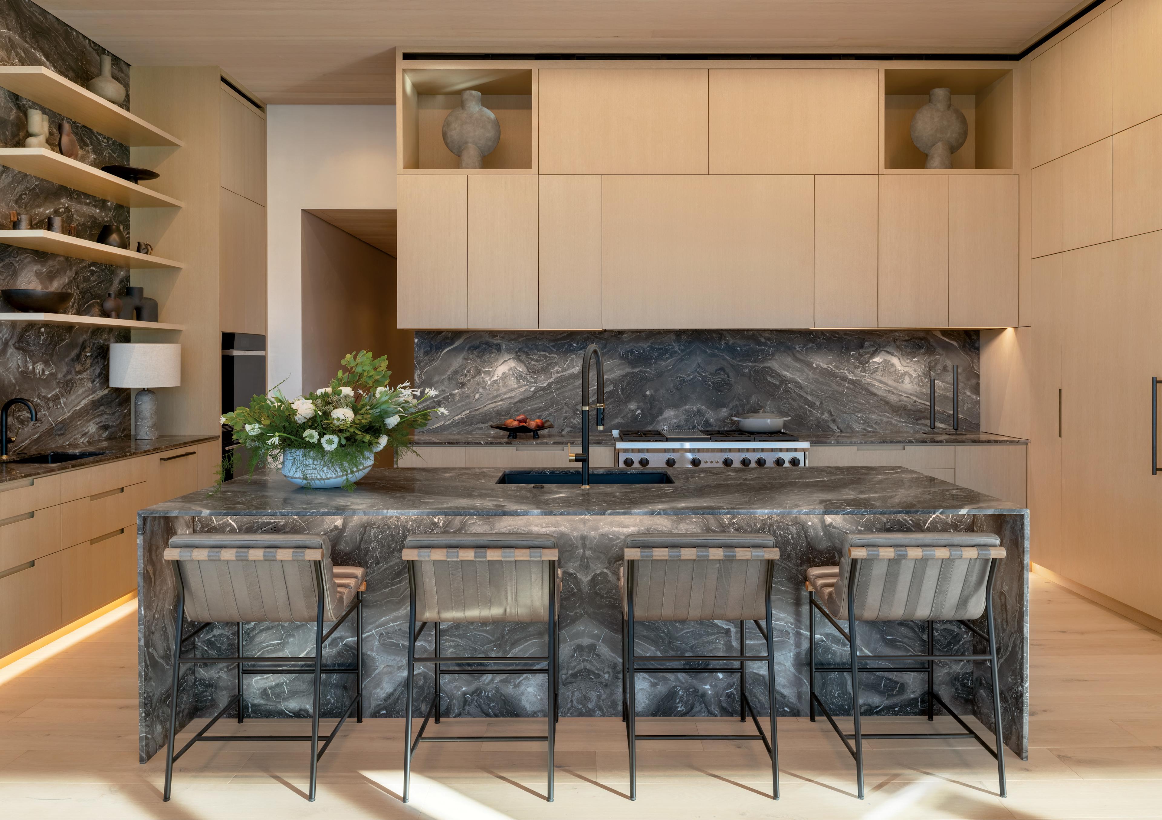 Modern kitchen with light wooden cabinets, dark marble countertops, and backsplash. Features an island with four chairs and a vase of flowers.