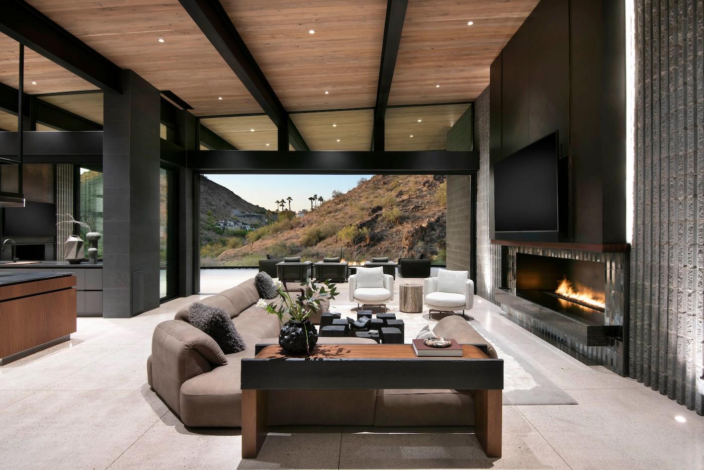 Modern living room with a cozy fireplace, large sofa, elegant decor, and floor-to-ceiling windows showcasing a scenic hillside view. Warm, inviting ambiance.